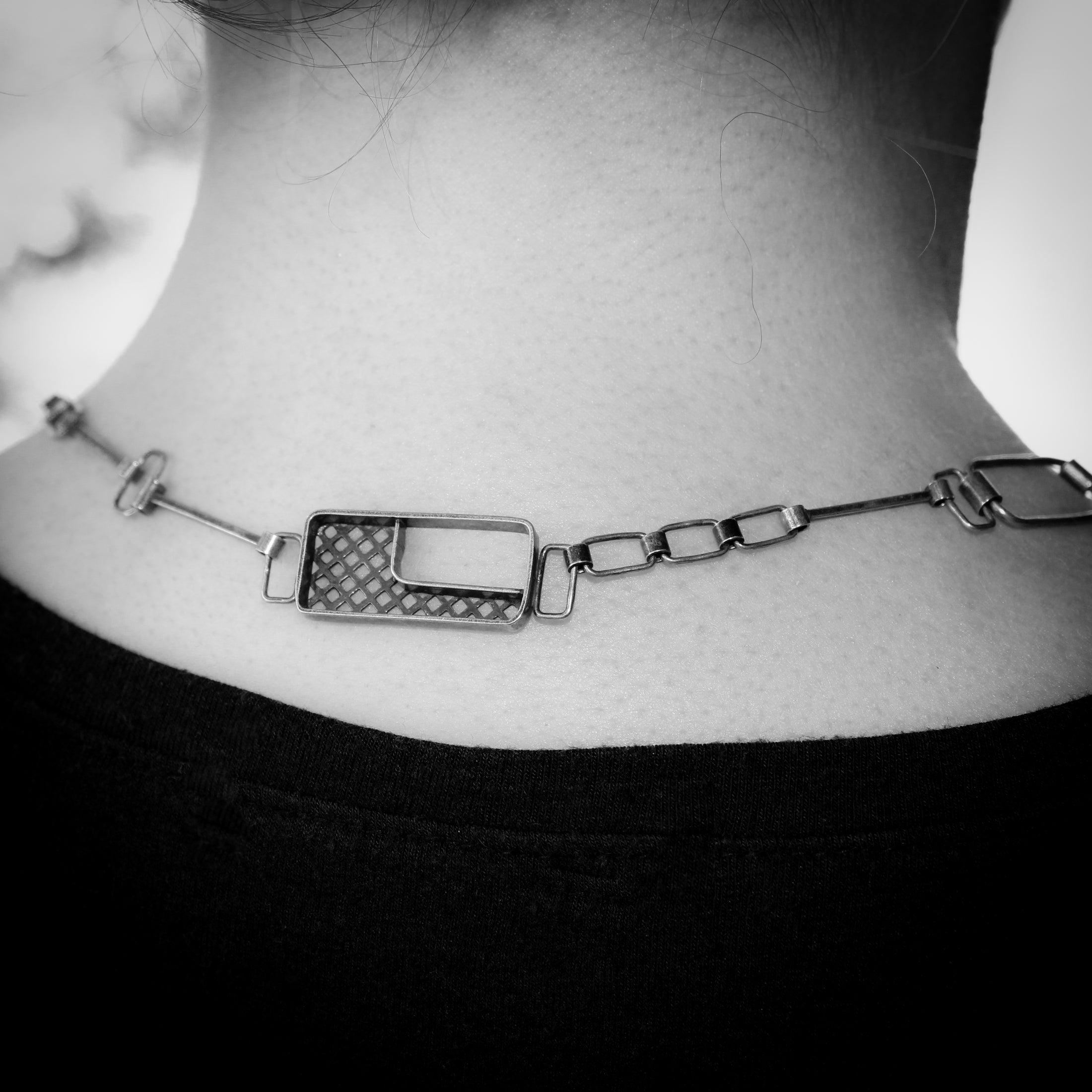 The black-and-white image shows the back of a person's neck adorned with a silver necklace featuring an additional small pendant that rests elegantly at the center. The pendant has a rectangular shape with an open geometric design, including a grid pattern. The chain itself consists of a mix of rectangular and square-shaped links, creating a minimalist, industrial aesthetic. This decorative pendant adds a unique touch to the back of the necklace, offering visual interest from behind. The person is wearing a