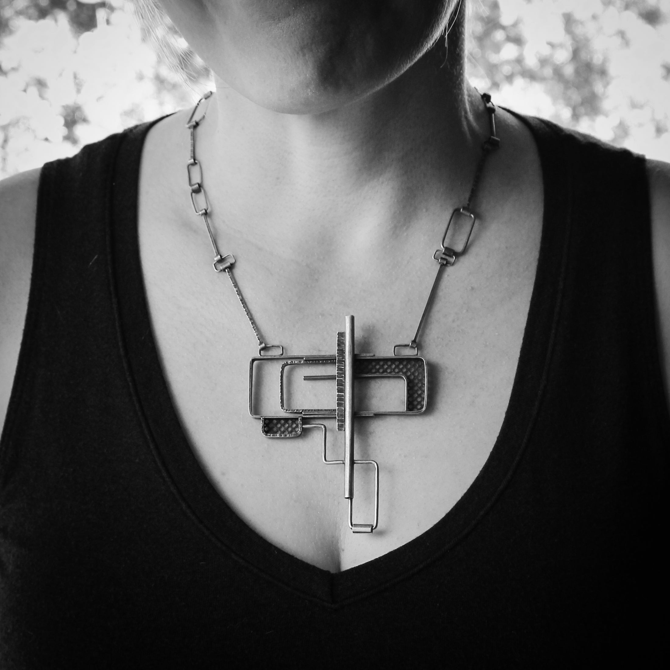 "Black and white image of a model wearing a sterling silver necklace featuring a bold geometric puzzle clasp pendant. The pendant has overlapping rectangular and square elements, with a mix of smooth and textured finishes. The chain consists of angular, rectangular links, matching the modern and architectural design of the necklace. The model is wearing a black top, and the pendant sits at the center of the chest.