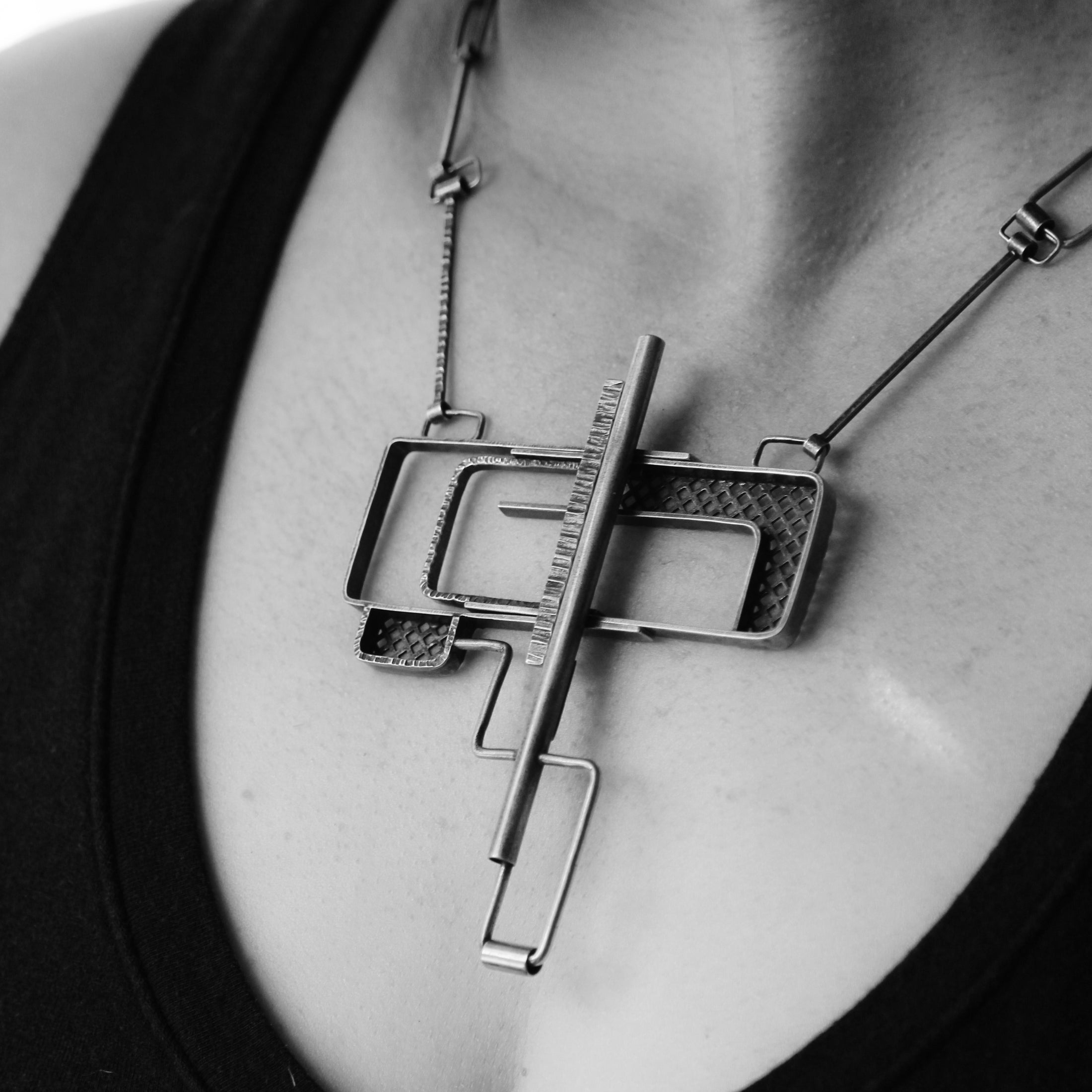 Close-up black and white image of a sterling silver puzzle clasp necklace worn by a model. The necklace features a complex geometric pendant with overlapping rectangular and square shapes, textured sections, and smooth metal bars, creating a modern and architectural design. The chain links are similarly angular and complement the pendant's bold structure.