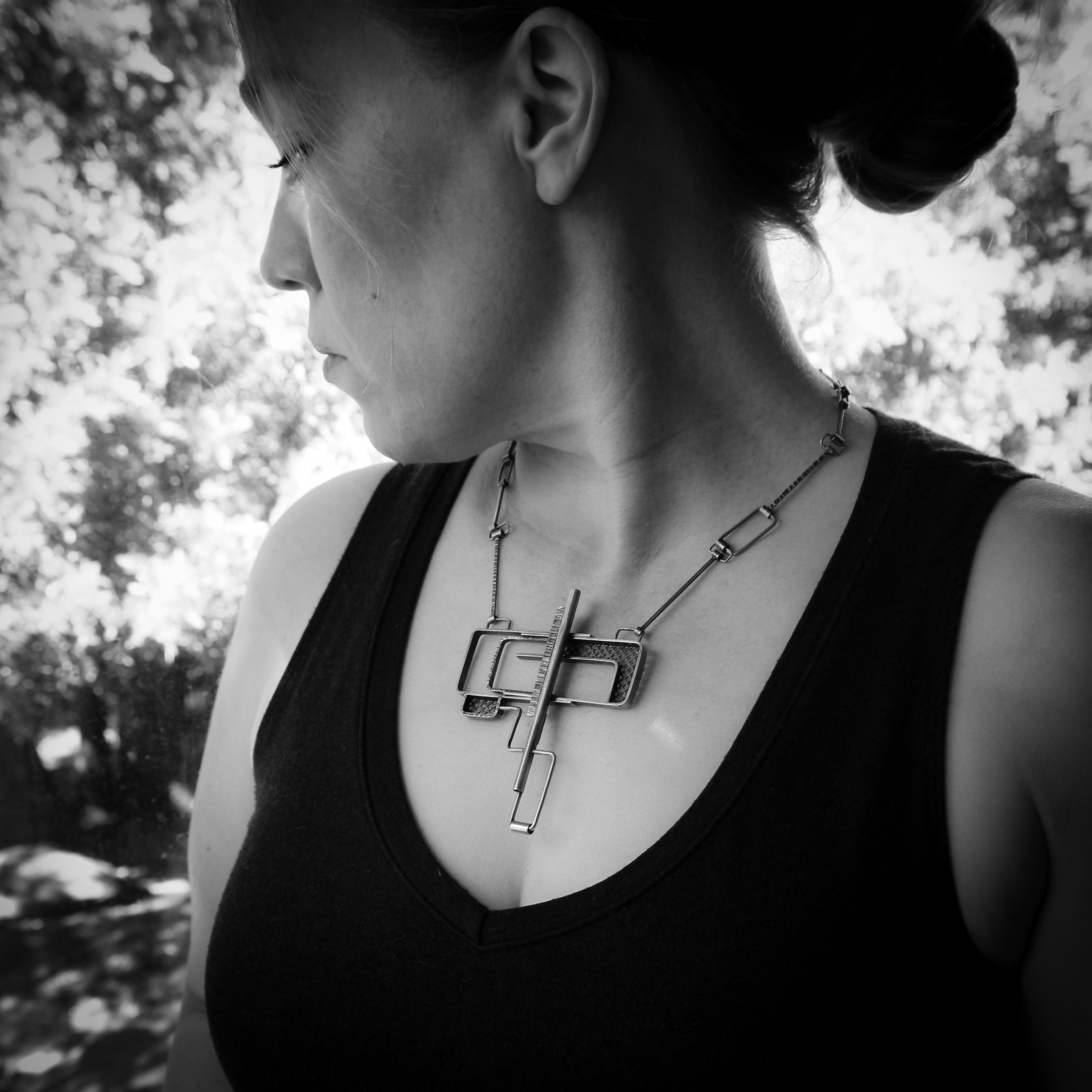 Black-and-white photo of a person wearing a geometric sterling silver pendant with an industrial design. The pendant features a combination of open rectangular shapes and mesh-textured sections, suspended from a chain with angular links. The person is shown in profile, wearing a sleeveless top, with the focus on the necklace. The background shows soft natural light filtering through trees, creating a serene and artistic atmosphere.