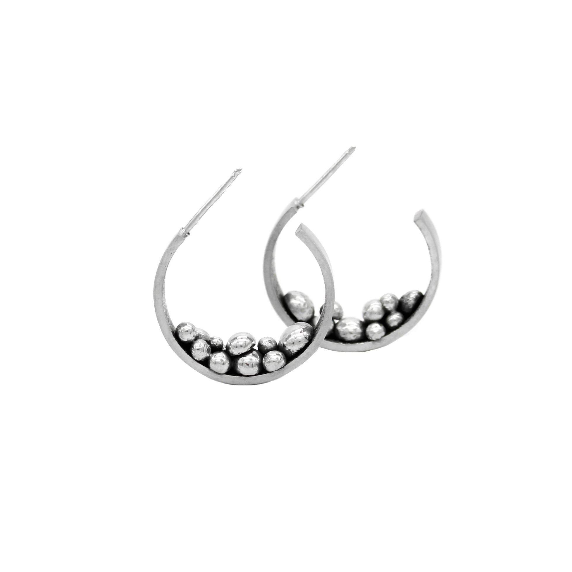 Two sterling silver, handmade earring hoops standing on their side, fabricated from 5 millimeter wide rectangle wire. Adorning the lower half of each hoop are assorted asymmetrical silver balls of varying balls nesting on top of each other. The interior of the hoops has a blackened finish, while the exterior is meticulously polished to a matte luster. 