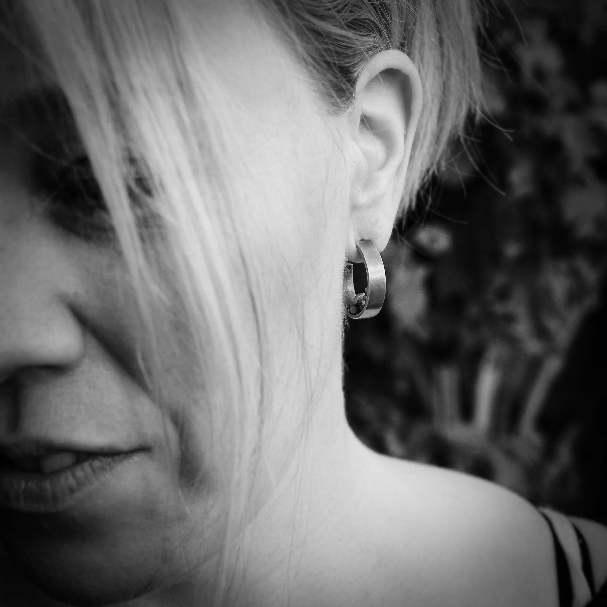 Black and white image of left half of woman's face, wearing a handmade sterling silver hoop, with silver balls on the interior, and a matte finish on the exterior. A blurred tree can be seen in the background. 