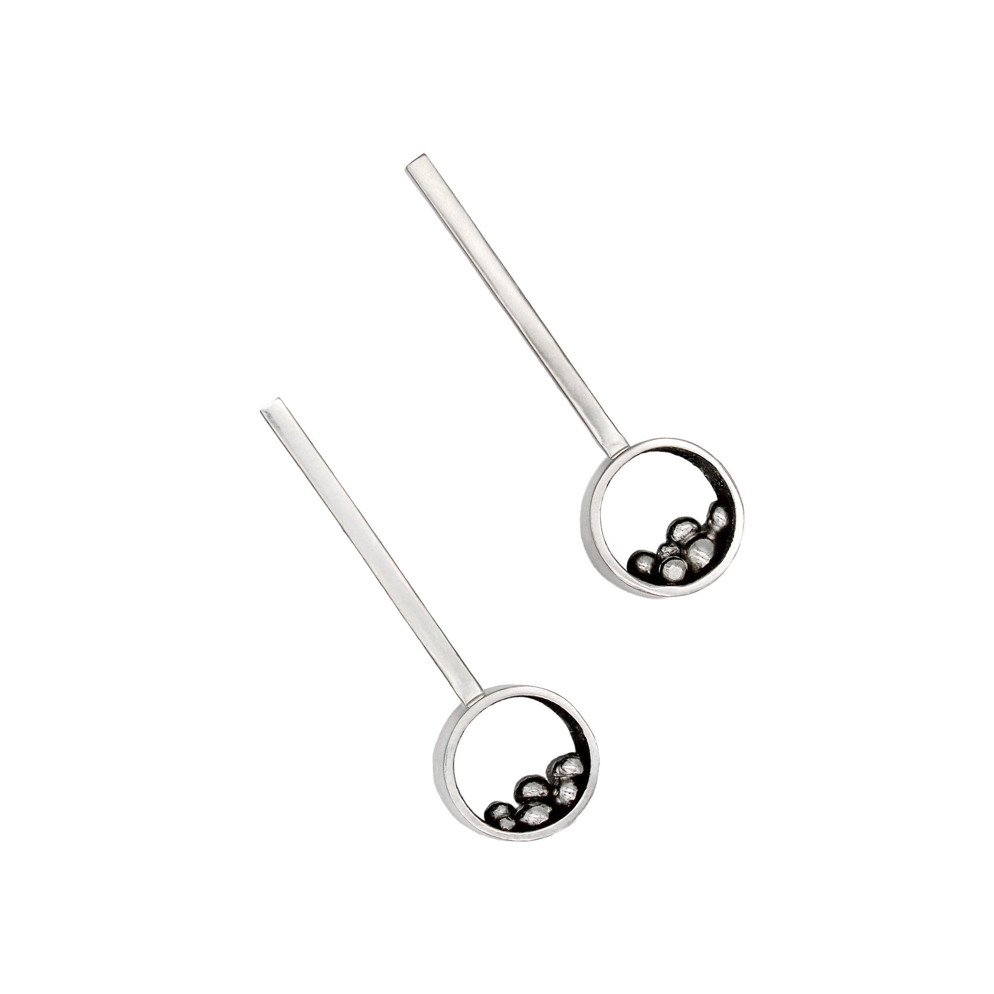 Pair of hand fabricated earrings in sterling silver, featuring a 10-millimeter circle connected to a 2-inch long rectangular bar with an ear post. The lower half of each circle has an array of asymmetrical silver balls of various sizes intricately nested. The circles' interiors showcase a sleek blackened and high polished finish, while the exteriors are a brushed matte luster. Both earrings laying on their back, and positioned next to each other on a white background.