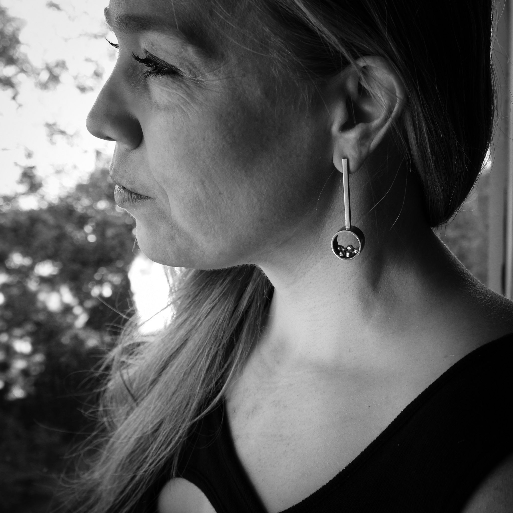 Black and white side profile of a woman's head featuring handmade sterling silver earrings. The earrings consist of a circular element, a rectangular bar, and assorted asymmetrical balls arranged in the circle's center. The woman is adorned in a black shirt, with blurred trees serving as the background.