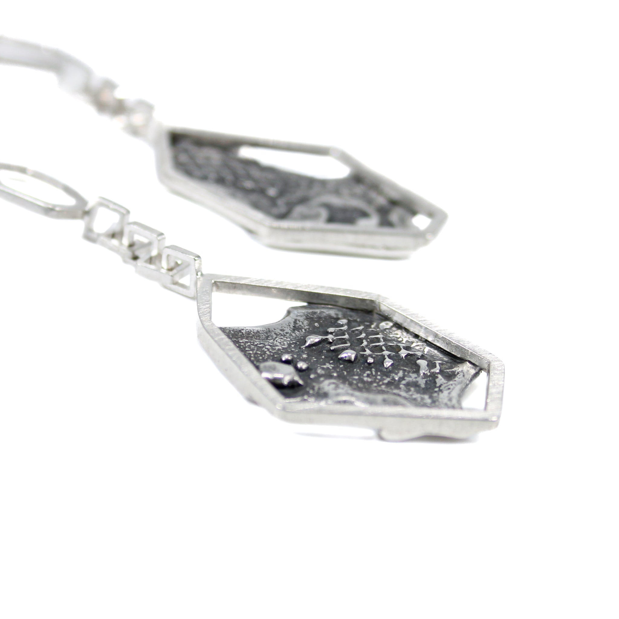 Close-up side view of blackened, brutalist inspired texture on backplate of earrings. Silver mesh, blobs and grit can be seen in the texture. The backplate is framed by an asymmetric hexagon, attached to some square links. The other earring is seen blurred in the background. 