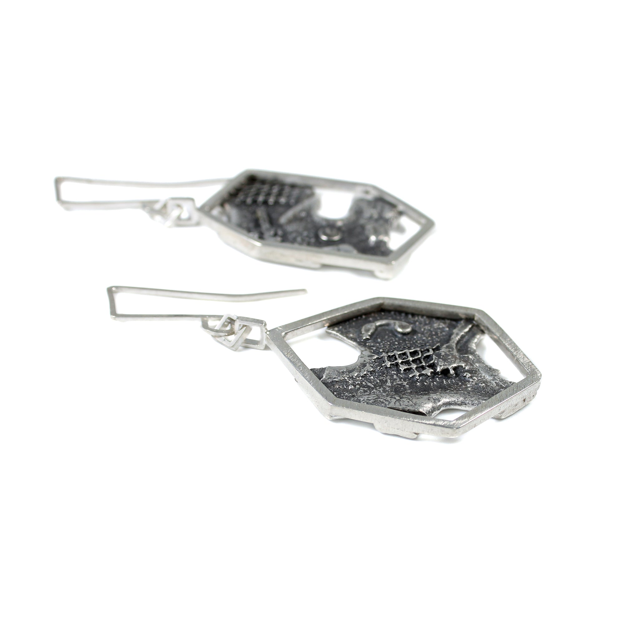 Close-up side view of blackened, brutalist inspired texture on backplate of earrings. Silver mesh, blobs and grit can be seen in the texture. The backplate is framed by an asymmetric hexagon, attached to some square links and a custom made square ear wire. The other earring is seen blurred in the background. 