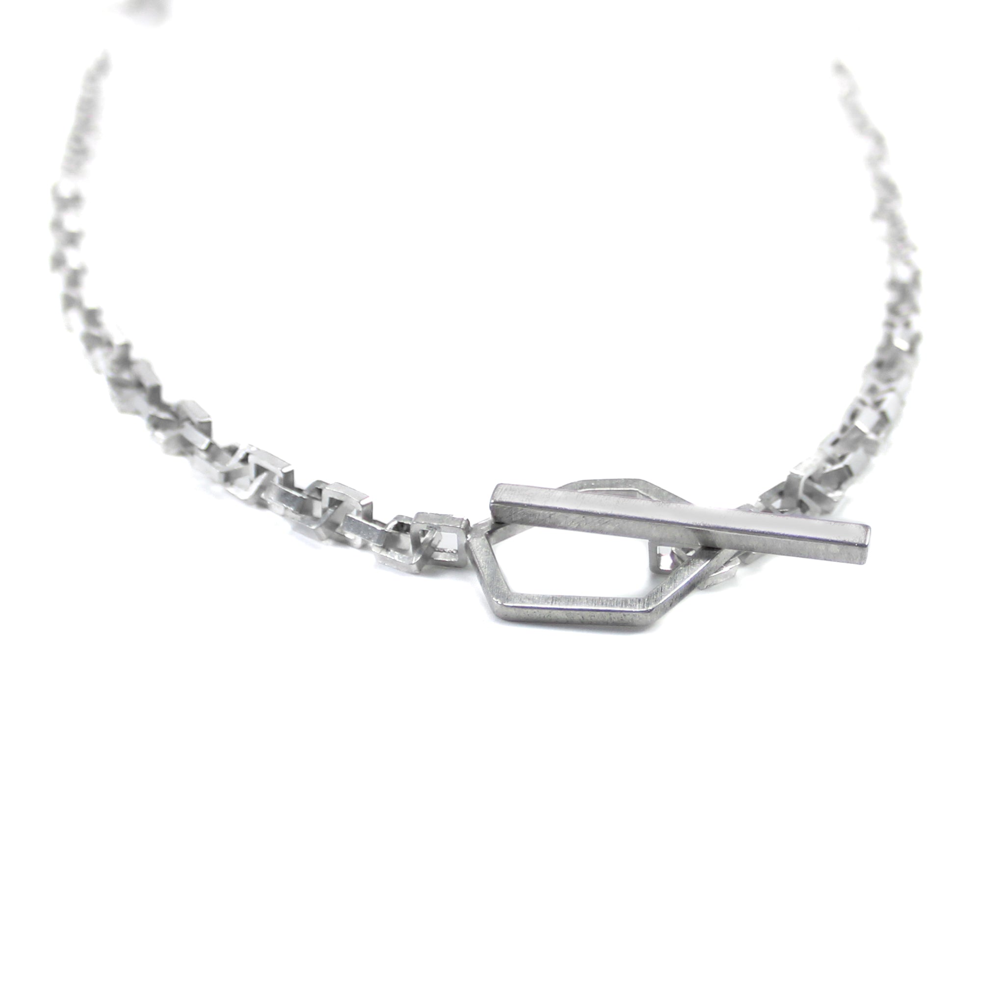 Close up of sterling silver toggle clasp, featuring an asymmetric, hexagon link attached to right-side of square link chain and a long rectangular bar attached to left-side of square link chain. The bar is threaded through the hexagon and shows a closed toggle clasp. 