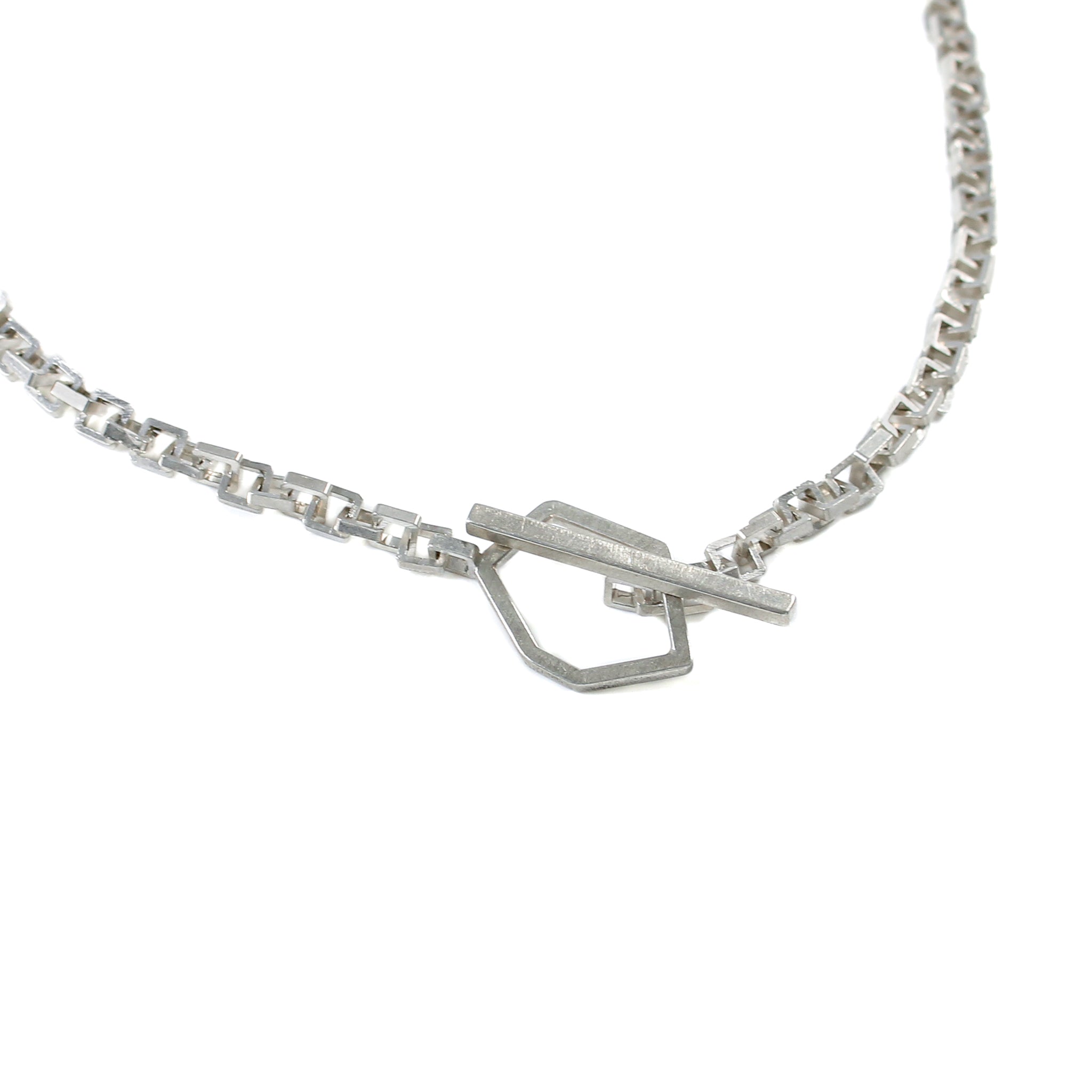 Close up of sterling silver toggle clasp, featuring an asymmetric, hexagon attached to right-side of square link chain and a rectangular bar attached to left-side of square link chain. The bar is threaded through the hexagon and shows a closed toggle clasp.