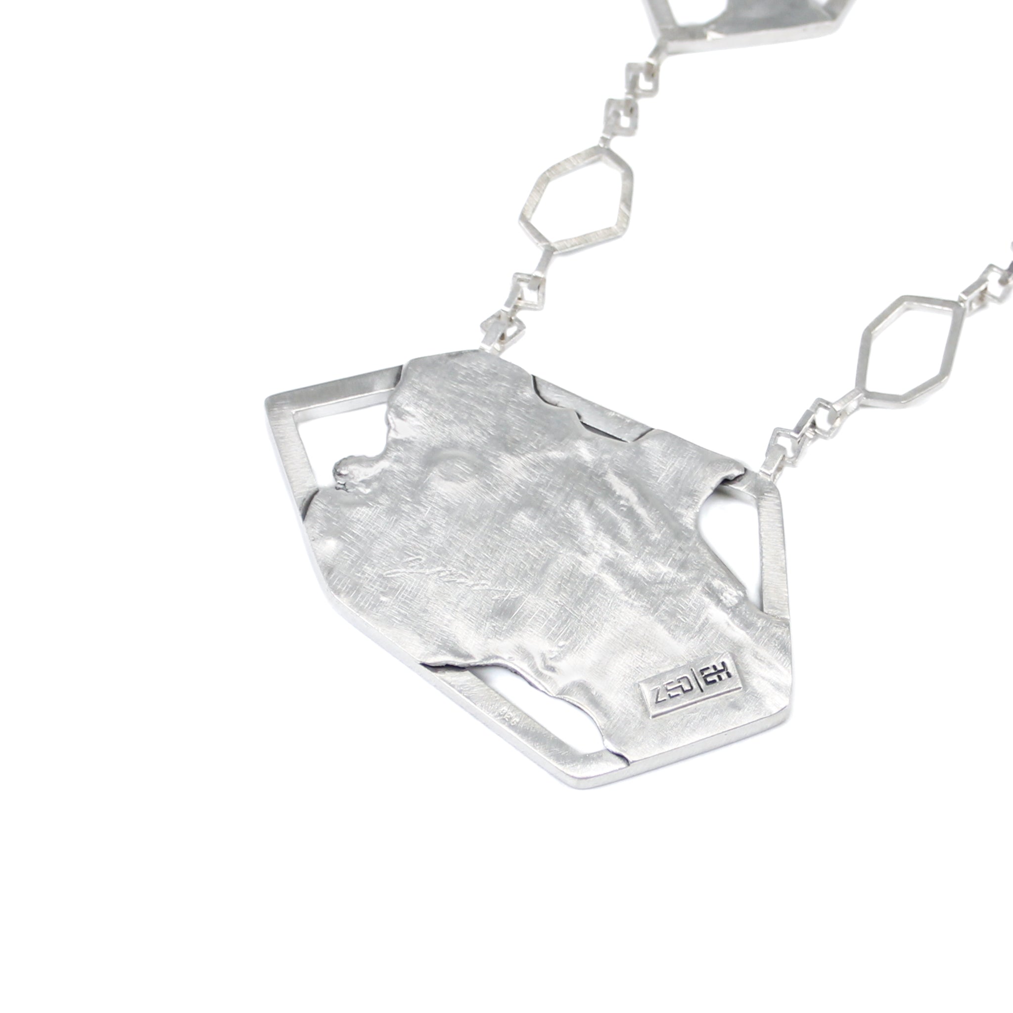 Back of asymmetric hexagonal pendant with a brushed matte sterling silver finish. Logo saying Zed Eh is stamped on edge of pendant, in black. Sqaure link chain attached to pendant is blurred in photo