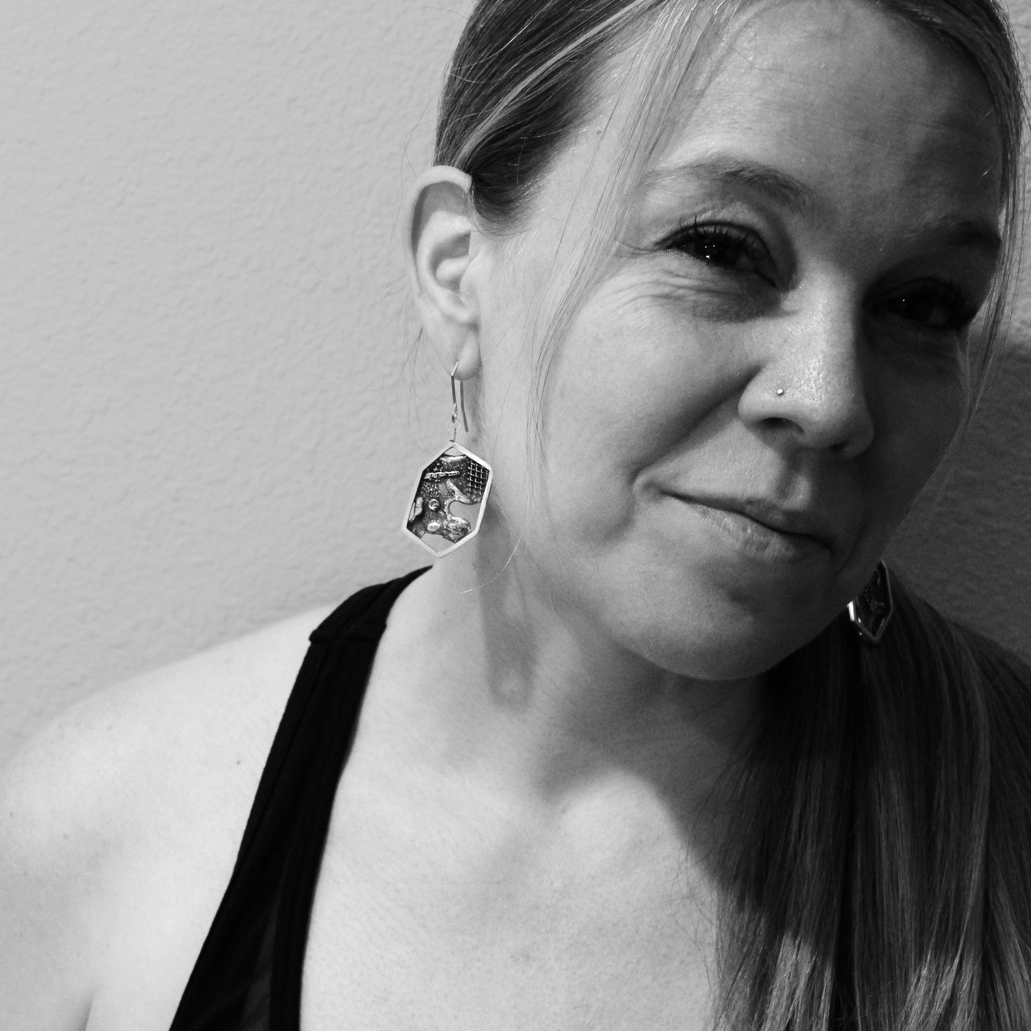 Black and white close-up of a woman's head, with a focus on the modern sterling silver hexagon earring in her right ear She wears a black tank top, sports a ponytail on her left side, and has a nose ring. She is looking off camera to the left.