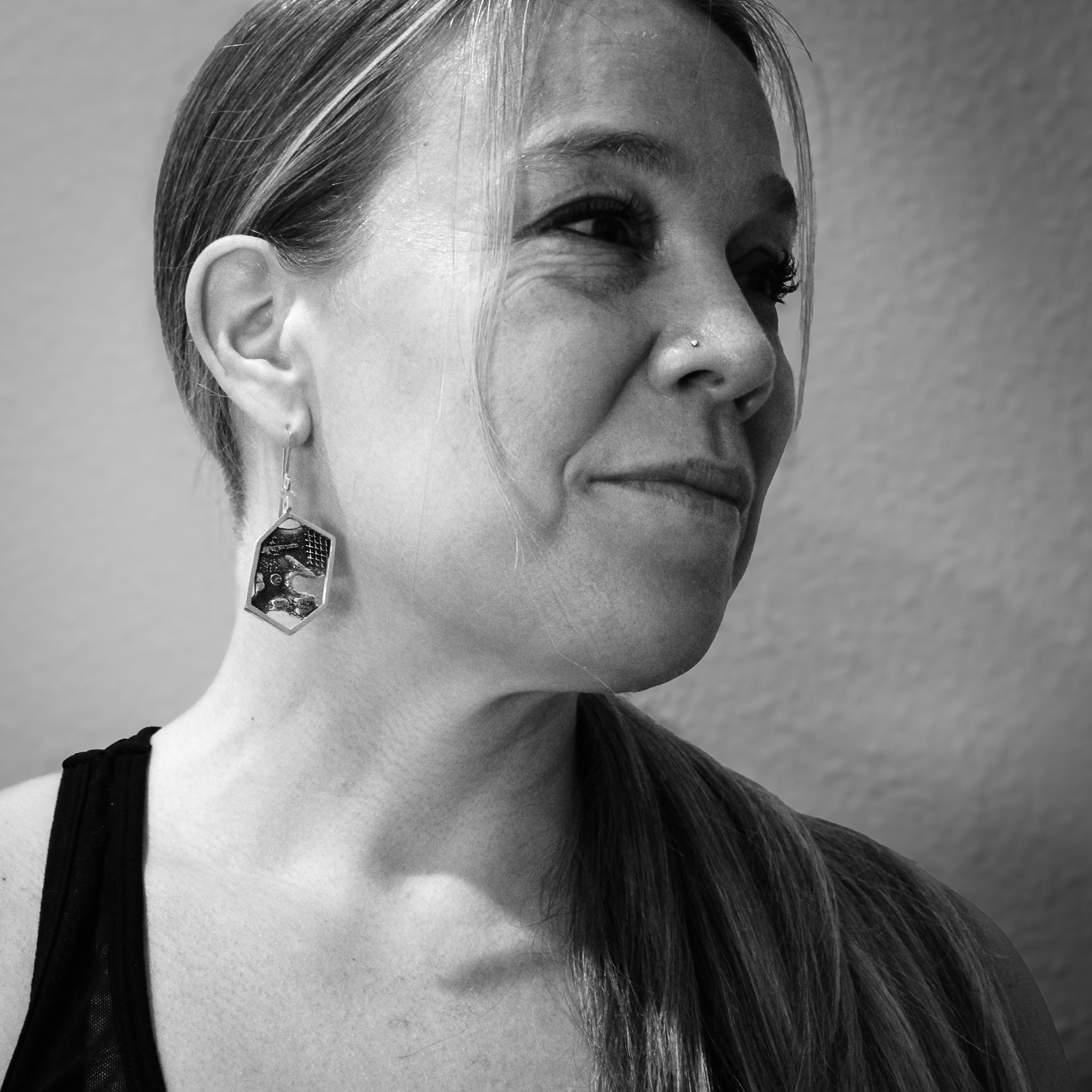 Black and white close-up of a woman's shoulders and head, with a focus on her modern sterling silver hexagon earrings. She wears a black tank top, sports a ponytail on her left side, and has a nose ring. Her head is tilted to the left and looks at the camera