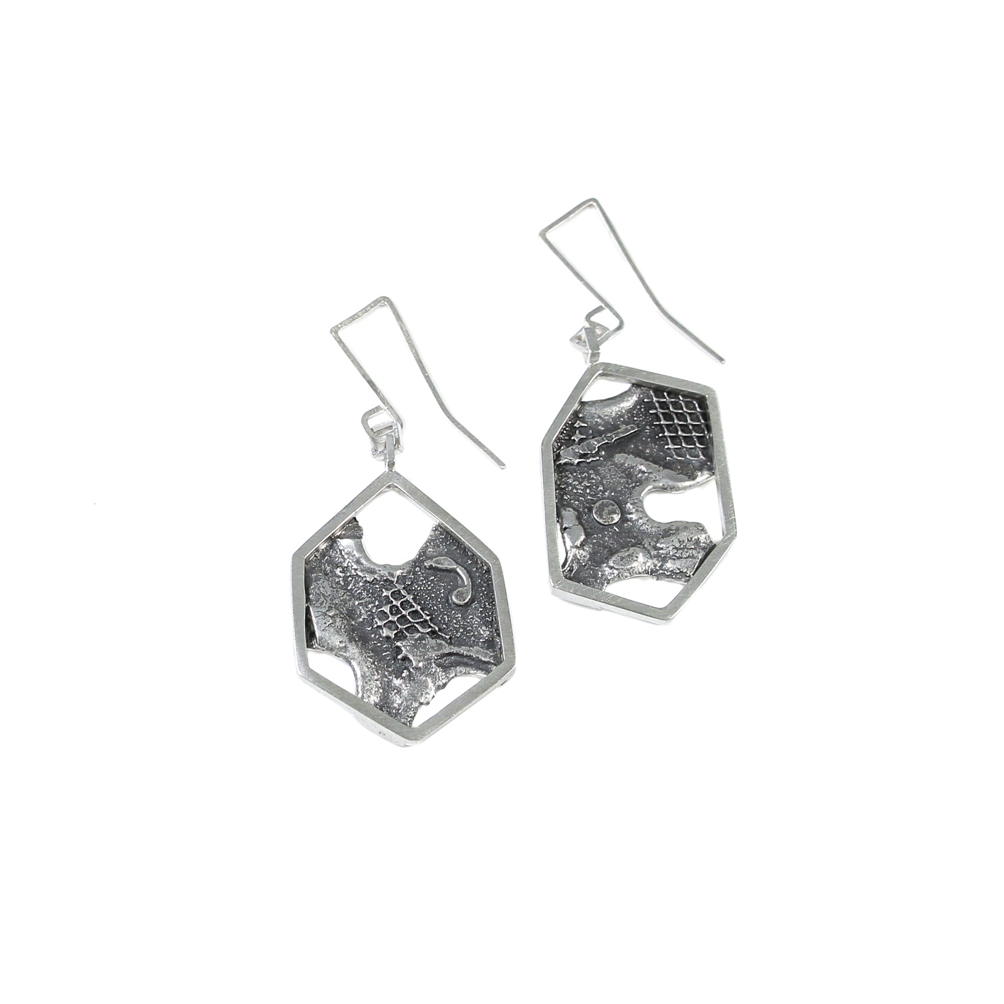 Sterling silver modern dangle earrings, on white background. Each earring has a large asymmetric hexagon, framing a blackened and brutalist textured backplate. This is attached to some square chain links finished with a custom made square shaped ear wire.