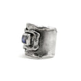 Load image into Gallery viewer, Side angle of sterling silver overlapping wrap ring, with melted edges and an oxidized rustic surface texture. A square, translucent, blue moonstone is roughly bezel set in the center of the ring. 
