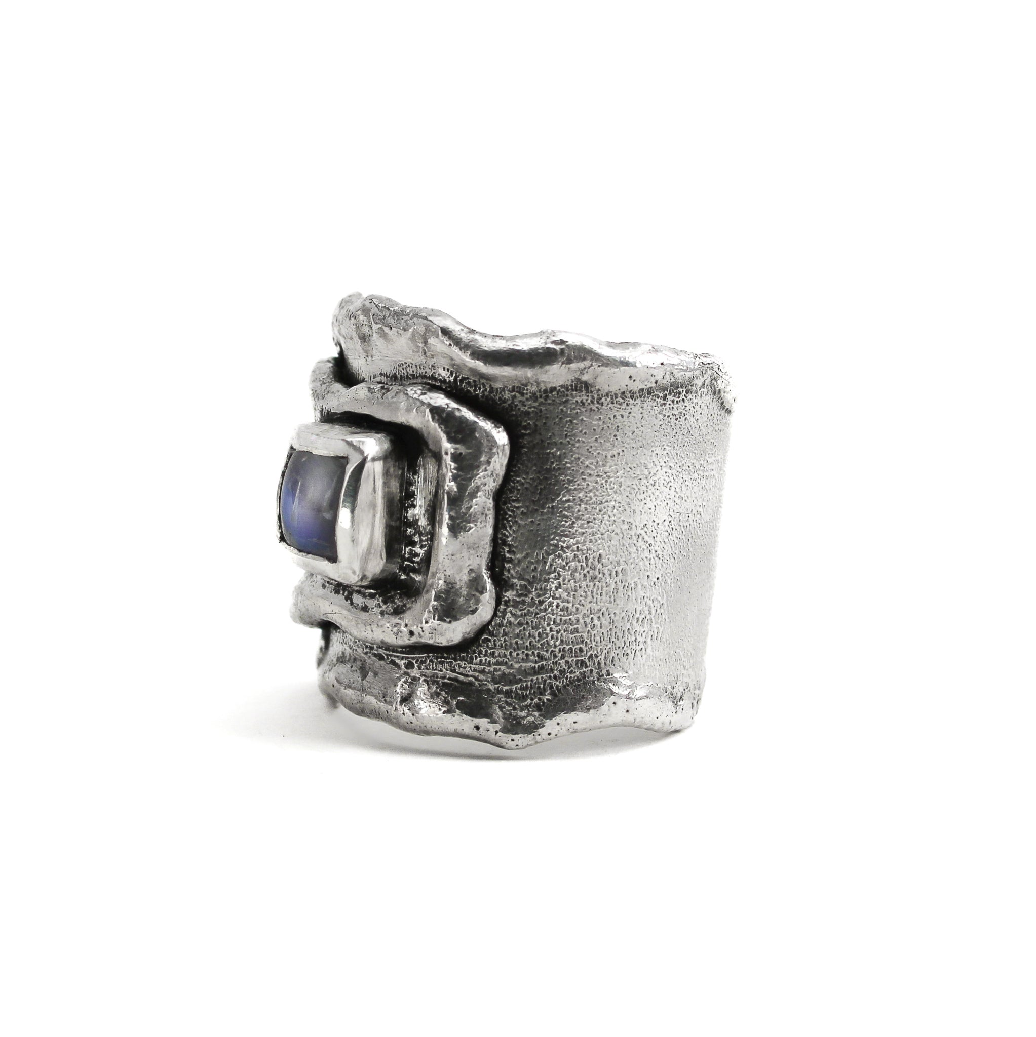 Side angle of sterling silver overlapping wrap ring, with melted edges and an oxidized rustic surface texture. A square, translucent, blue moonstone is roughly bezel set in the center of the ring. 