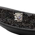 Load image into Gallery viewer, Sterling silver overlapping wrap ring with melted edges and an oxidized rustic surface texture, in  black dish with black pebbles. A square, translucent, blue moonstone is roughly bezel set in the center of the ring.
