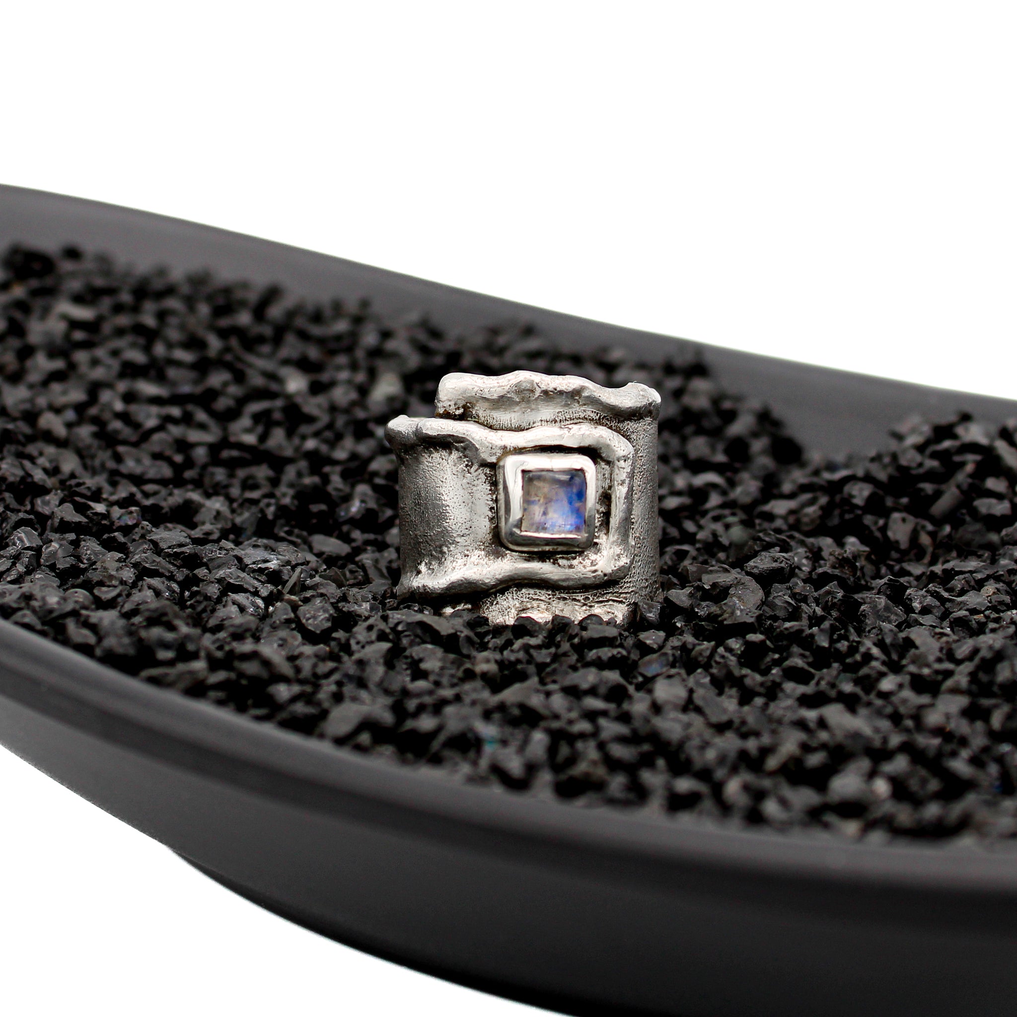 Sterling silver overlapping wrap ring with melted edges and an oxidized rustic surface texture, in  black dish with black pebbles. A square, translucent, blue moonstone is roughly bezel set in the center of the ring.