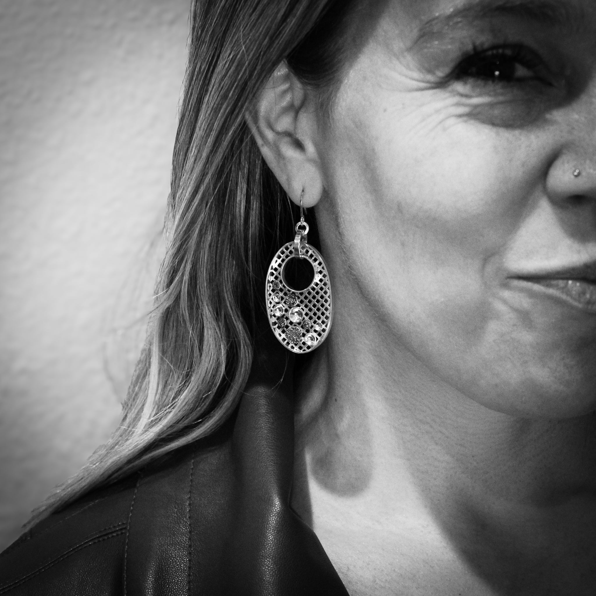 Black and white close-up of the right side of a woman's face, smiling and looking at the camera. The photo bisects her face down the middle. She wears a black leather jacket, and her hair is down, tucked behind her ear. The focal point is the handmade sterling silver and gold oval dangle earrings.