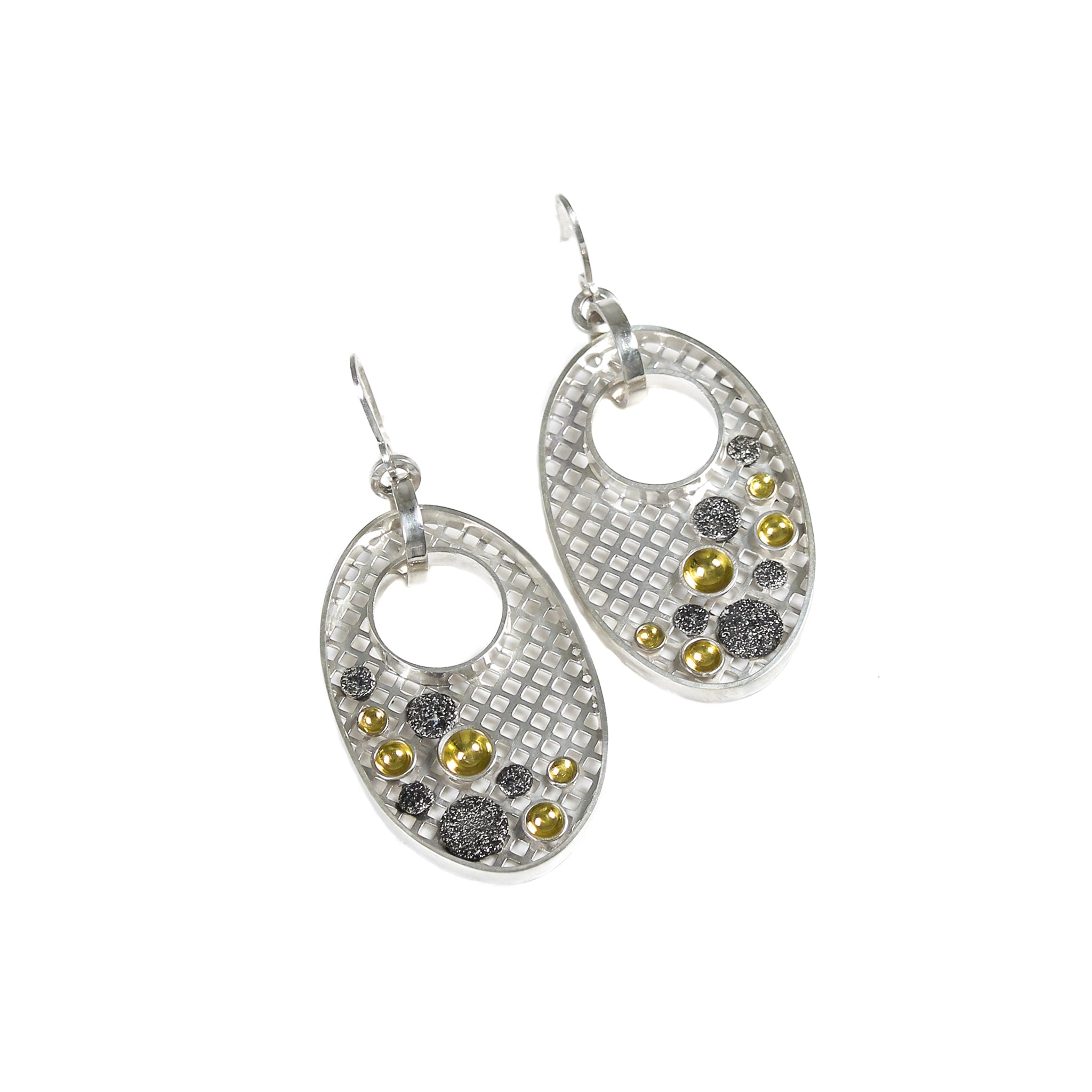 Sterling silver earrings with a contemporary design, featuring an oval shape. The back of the oval is covered with a silver mesh. A 10 millimeter circular hole is cut into the mesh near the top of the oval, allowing a wide silver hoop to weave through and link to a custom crafted ear hook. The oval mesh is embellished with 5 black discs and 5 gold cups of assorted sizes, scattered along the bottom and sides of the oval. Earrings are laying flat, on a white background.