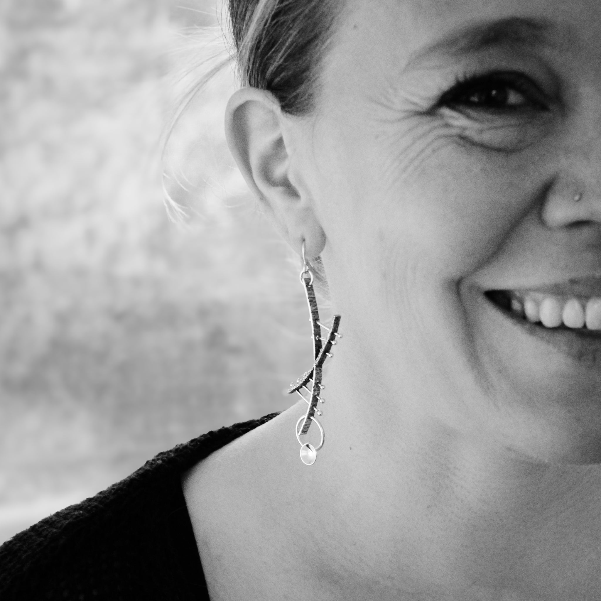 Black and white close-up of the right side of a woman's face, with her face bisected at the edge of the photo. She looks directly at the camera with a big smile. Wearing a black jacket with a large collar, the focus is on the modern silver and gold helix earrings she has on