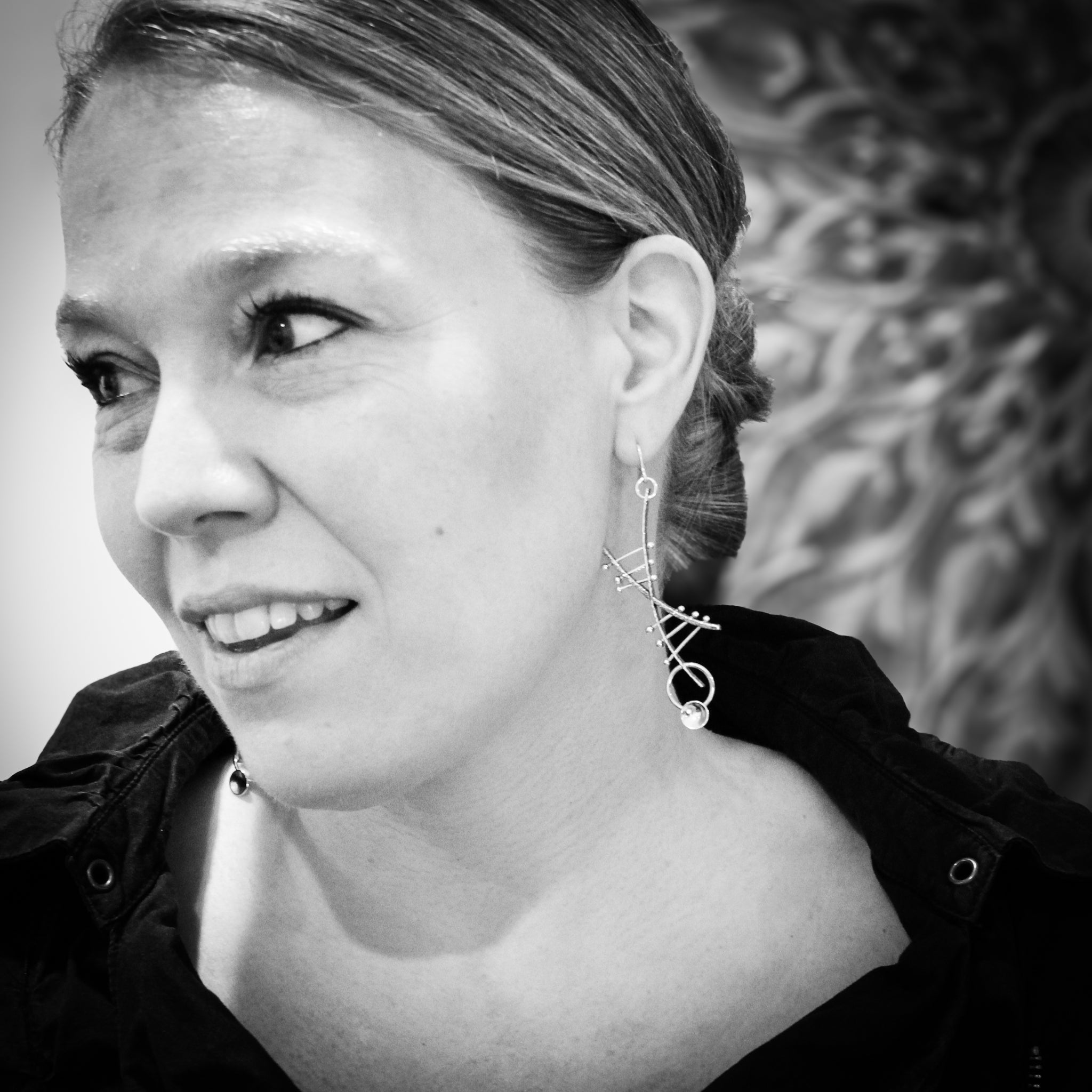 Black and white close-up of a woman's face and neck. She gazes off-camera to the right with a slight smile, her hair styled in a bun. Wearing a black jacket with a large collar, the focus is on the modern silver and gold helix earrings she is has on.