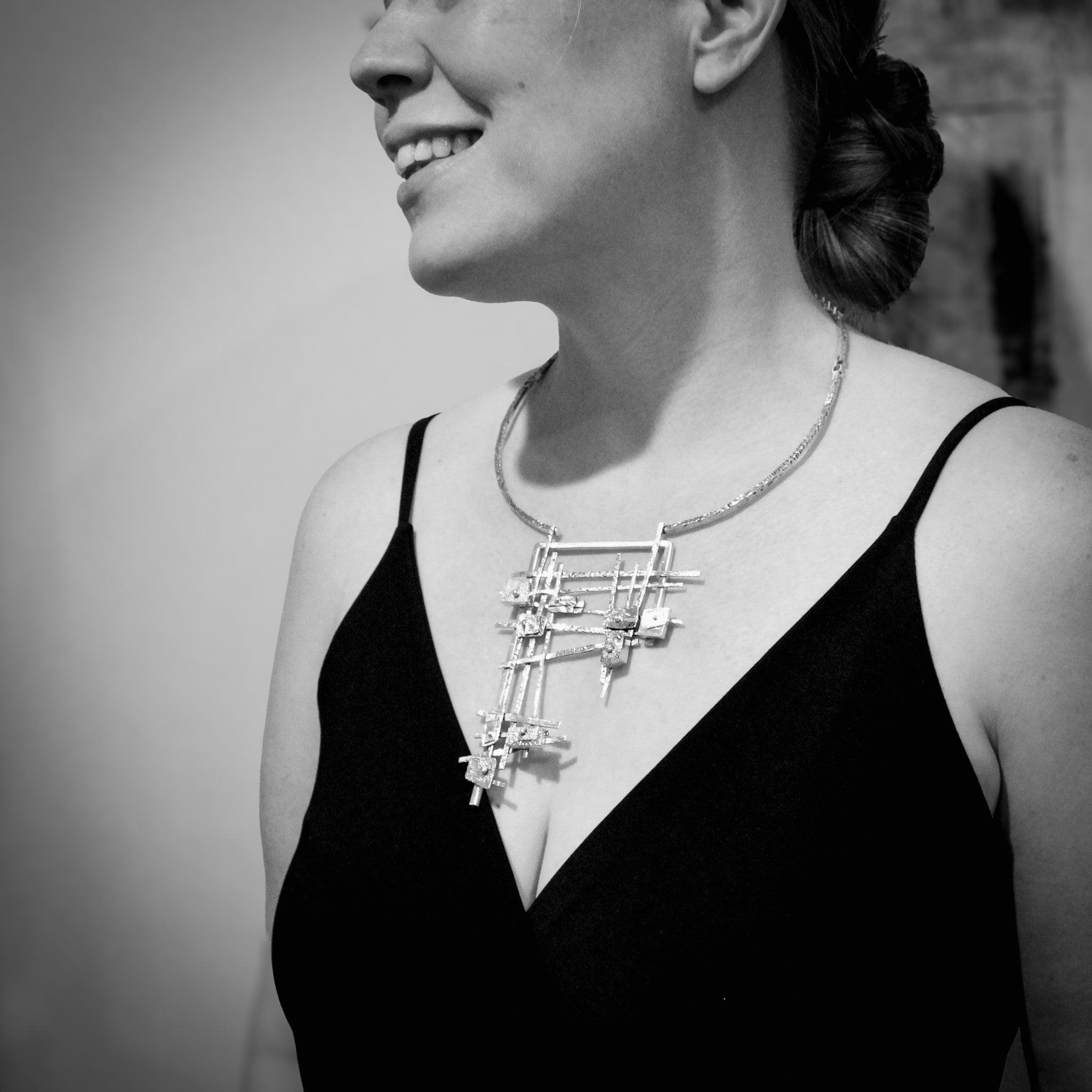 Black and white of a woman's torso, wearing a black spaghetti strap shirt. She smiles and gazes off-camera to the right. The focal point is the contemporary statement necklace she wears – a sterling neck collar adorned with a silver and gold geometric grid pendant.