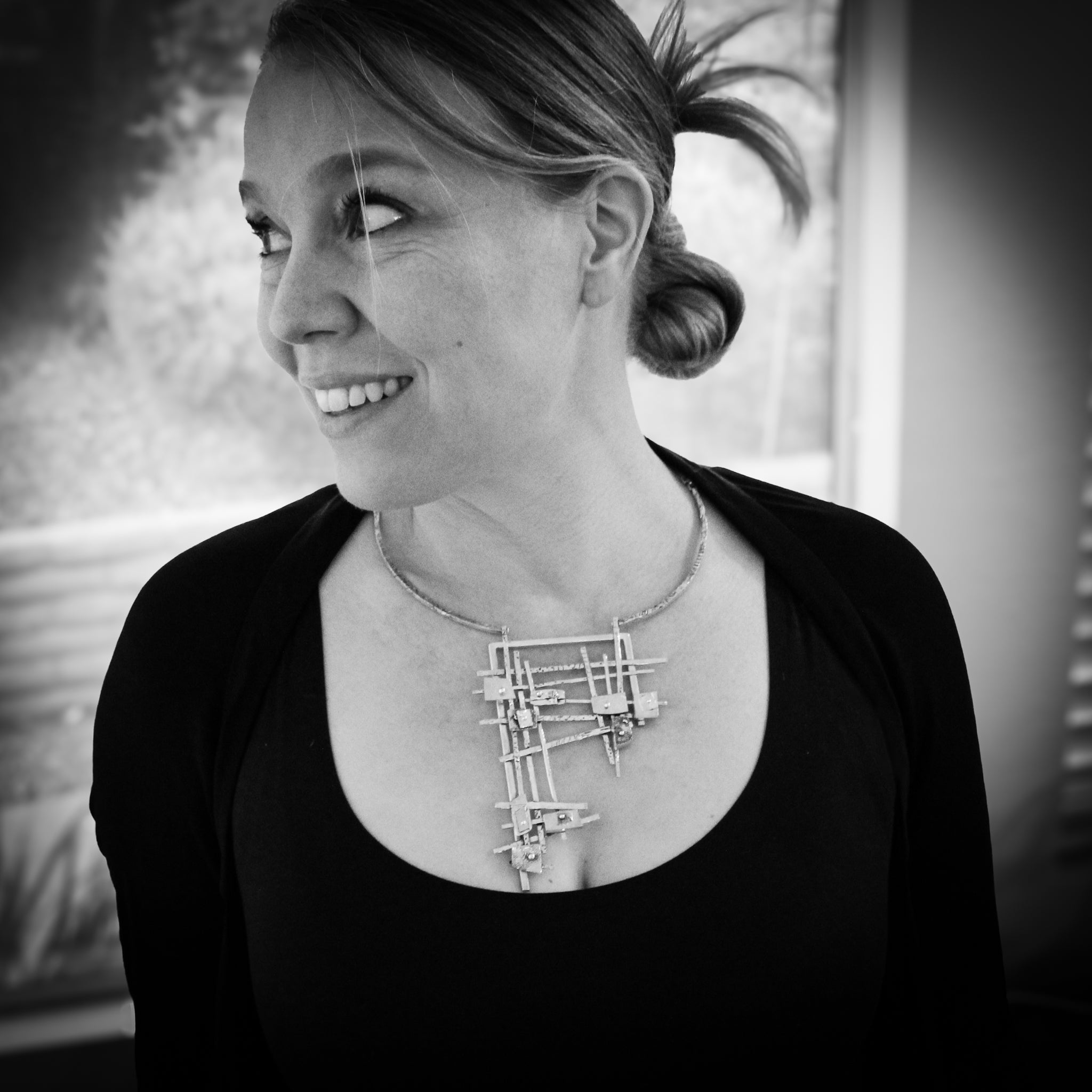 Black and white close up of a woman's torso and face. She is wearing a black long sleeve shirt, and is looking off camera to the right, with a smile on her face. The focal point is the statement necklace she wears – a sterling neck collar adorned with a silver and gold geometric grid.