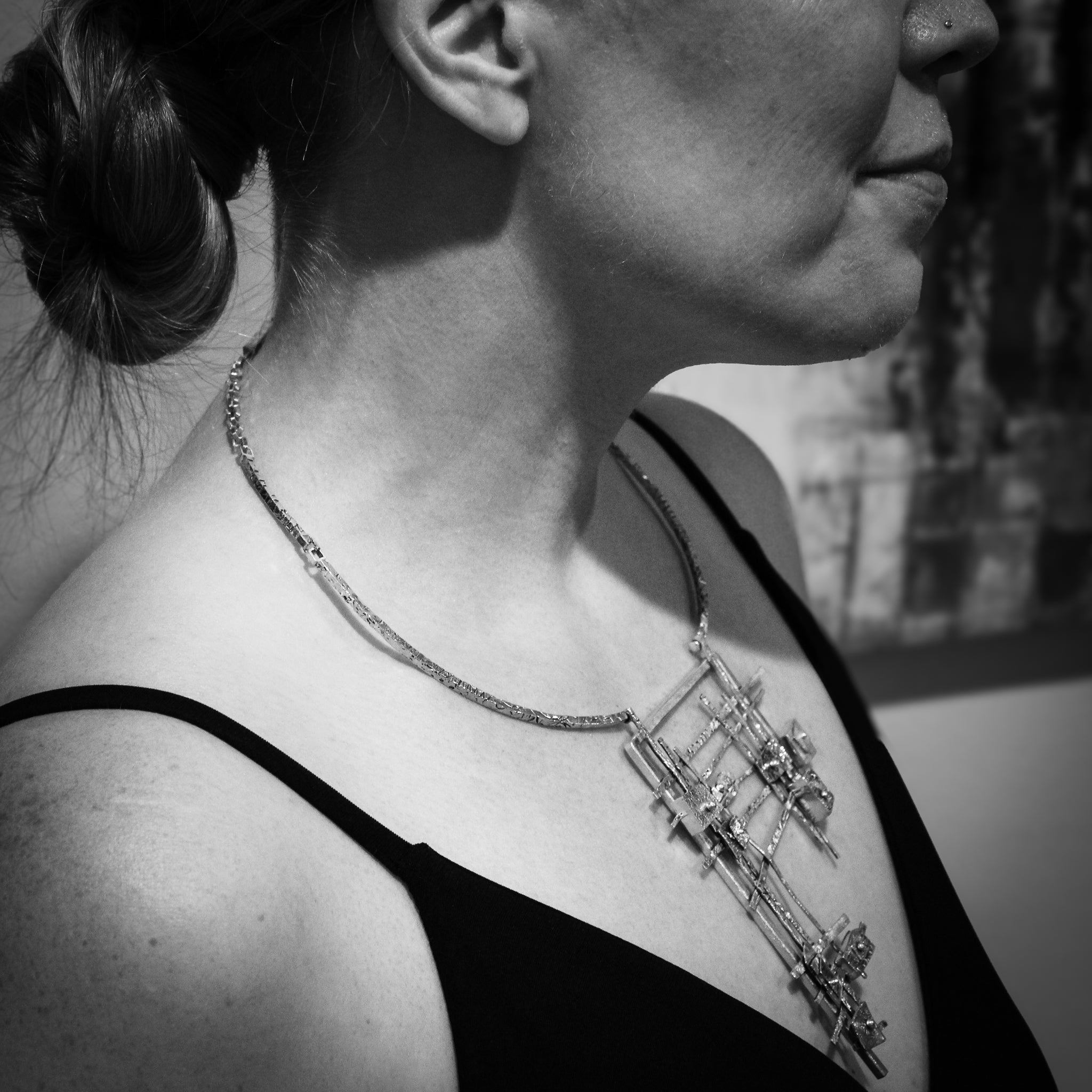 Black and white of a woman's torso, wearing a black spaghetti strap shirt. She smiles and gazes off-camera to the left. The focal point is a side view of the significant art necklace she wears – a sterling neck collar adorned with a silver and gold geometric grid pendant.