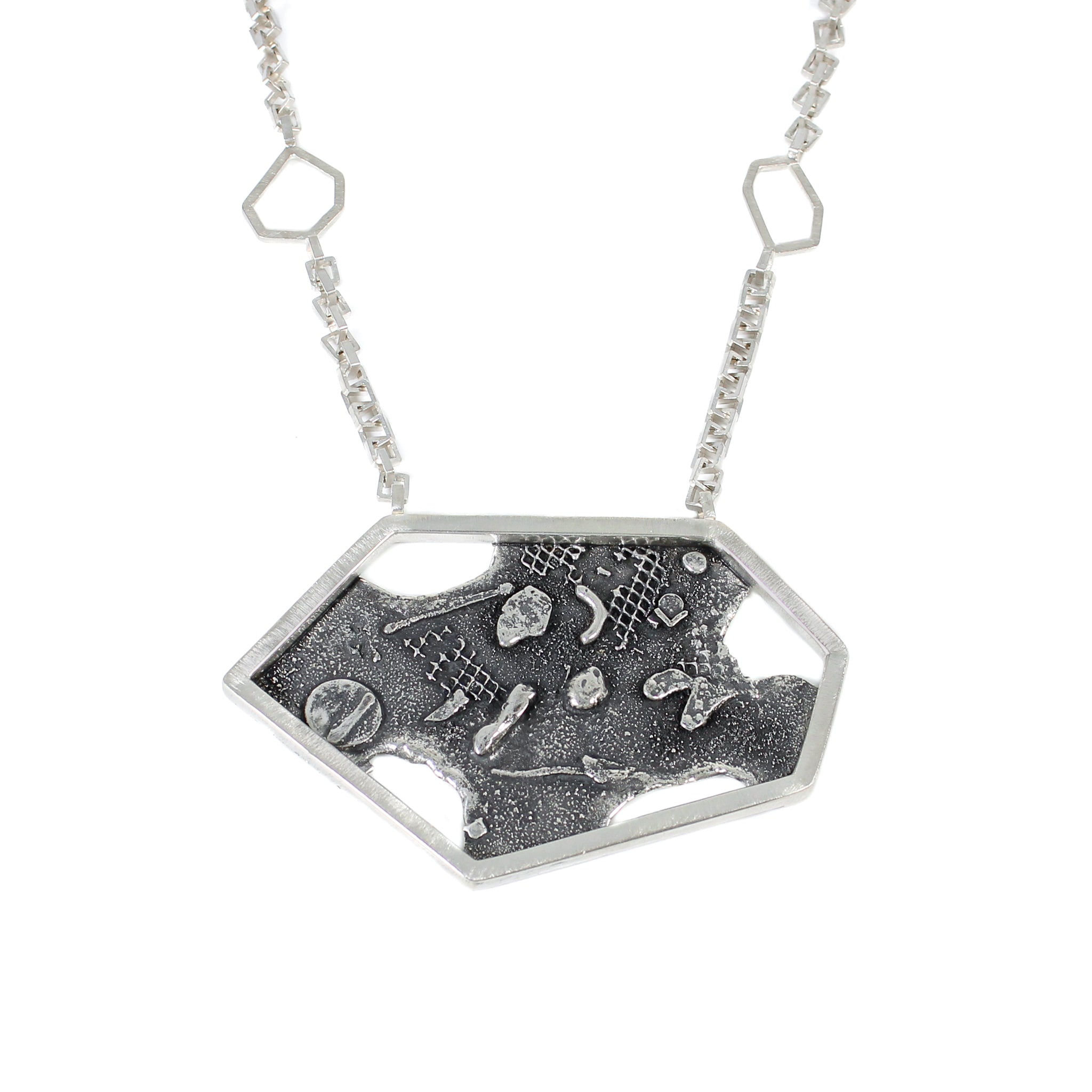 A close up, front view of a modern sterling silver pendant, featuring a large, asymmetric hexagon frame over a blackened, fused and brutalist textured blackplate. There are 4 melted holes in the backplate. The pendant is hung from a square link in link chain, with a small open hexagon 1.5" up the chain on either side.