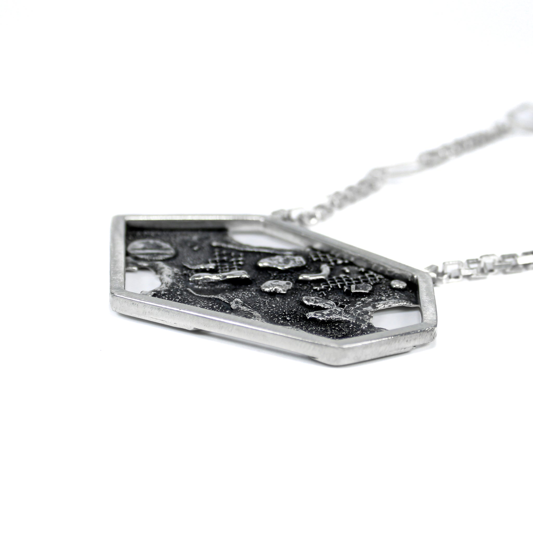 Laying flat side view of a contemporary sterling silver pendant, showing the right, outside edge of an asymmetric hexagon, made of thick square wire. The blackened backplate is slightly visible. The pendant is attached to a square link chain, that is blurred to view.