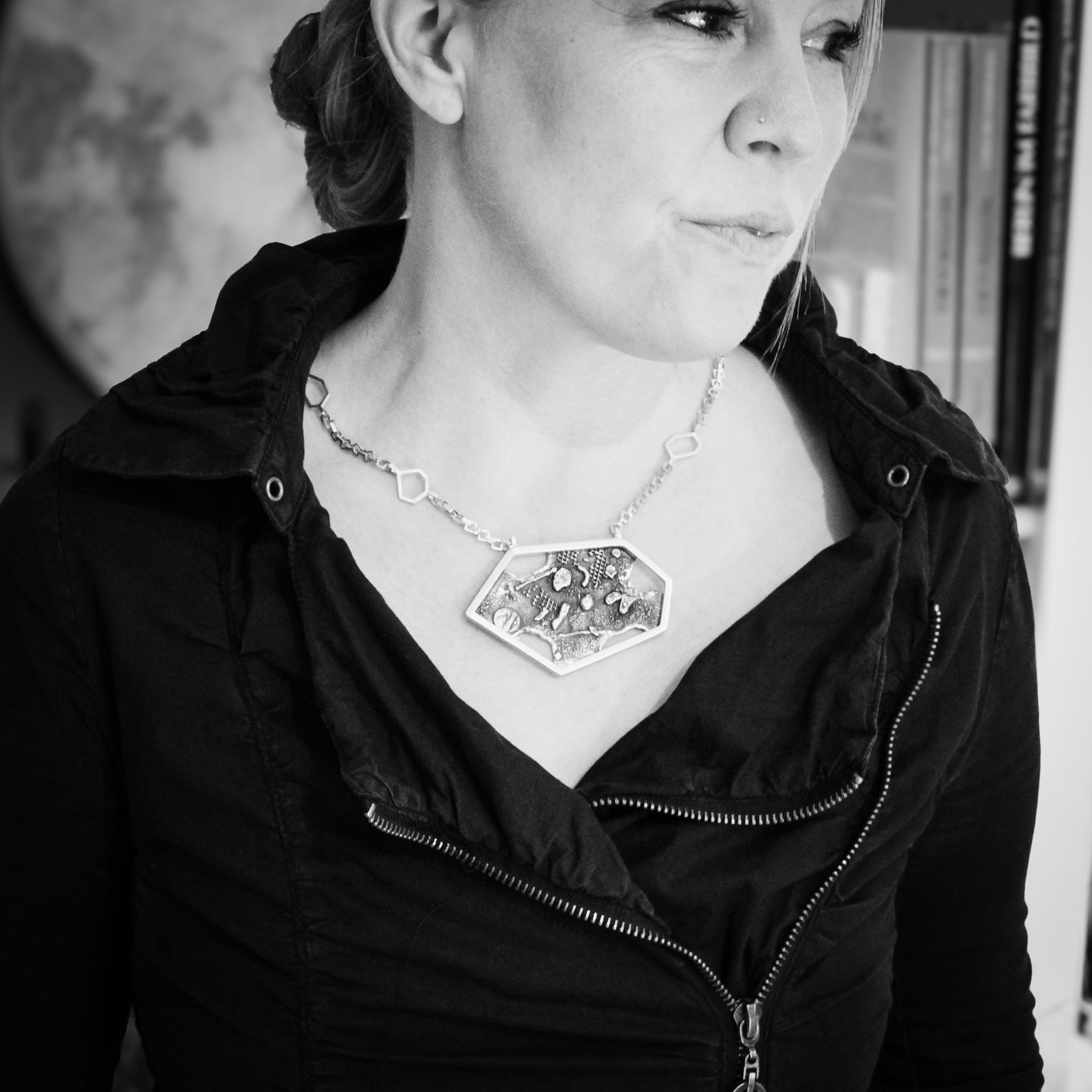 Black and white close-up of a woman's torso and head as she looks to the left. She wears a black jacket with two front zippers and a large collar, her hair styled in a loose bun. The focal point is the sterling silver modern hexagon pendant she is wearing. In the background there is a blurred bookshelf.