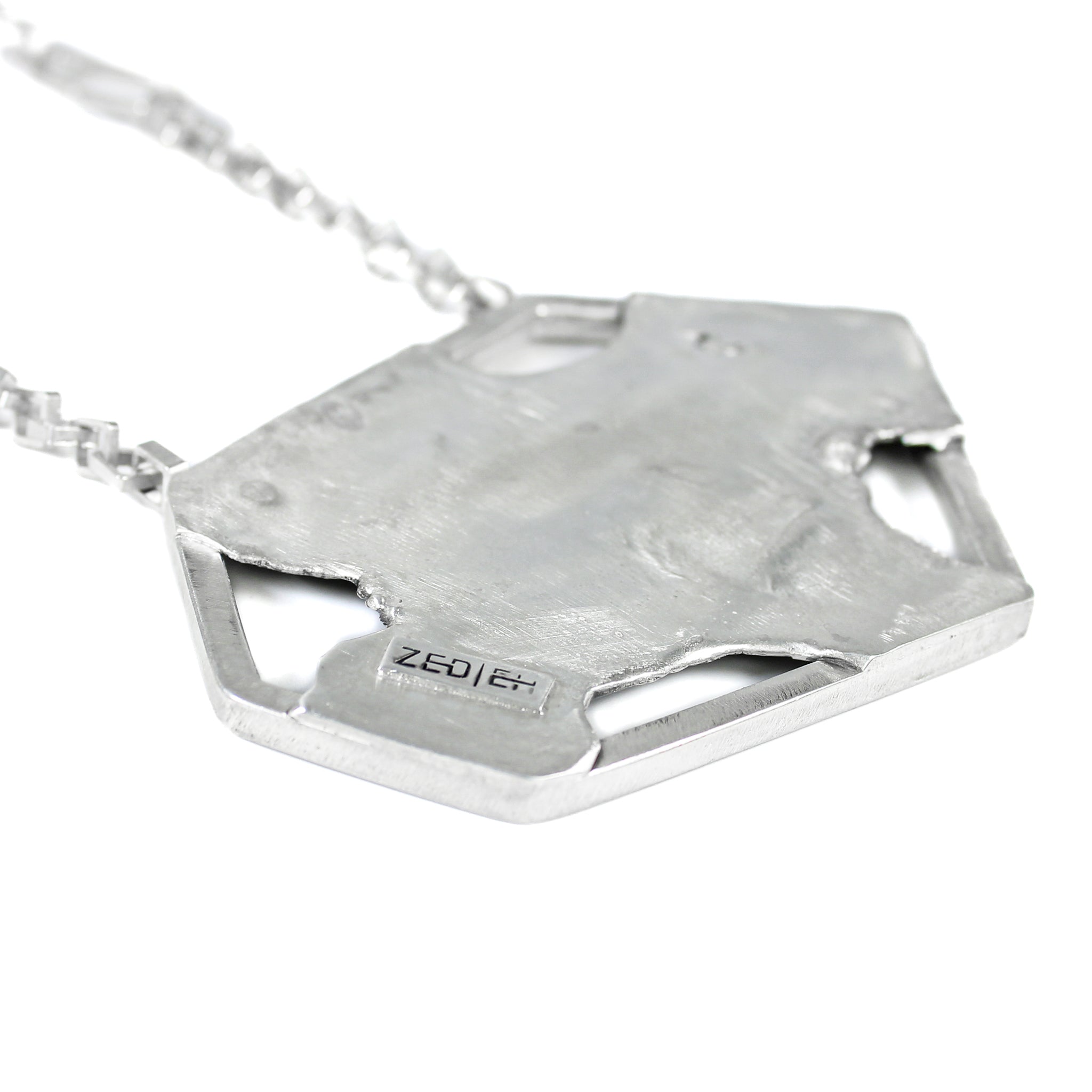 Back of asymmetric hexagonal pendant with a brushed matte sterling silver finish. Logo saying Zed Eh is stamped on edge of pendant, in black. Square link chain attached to pendant is blurred, on a white background