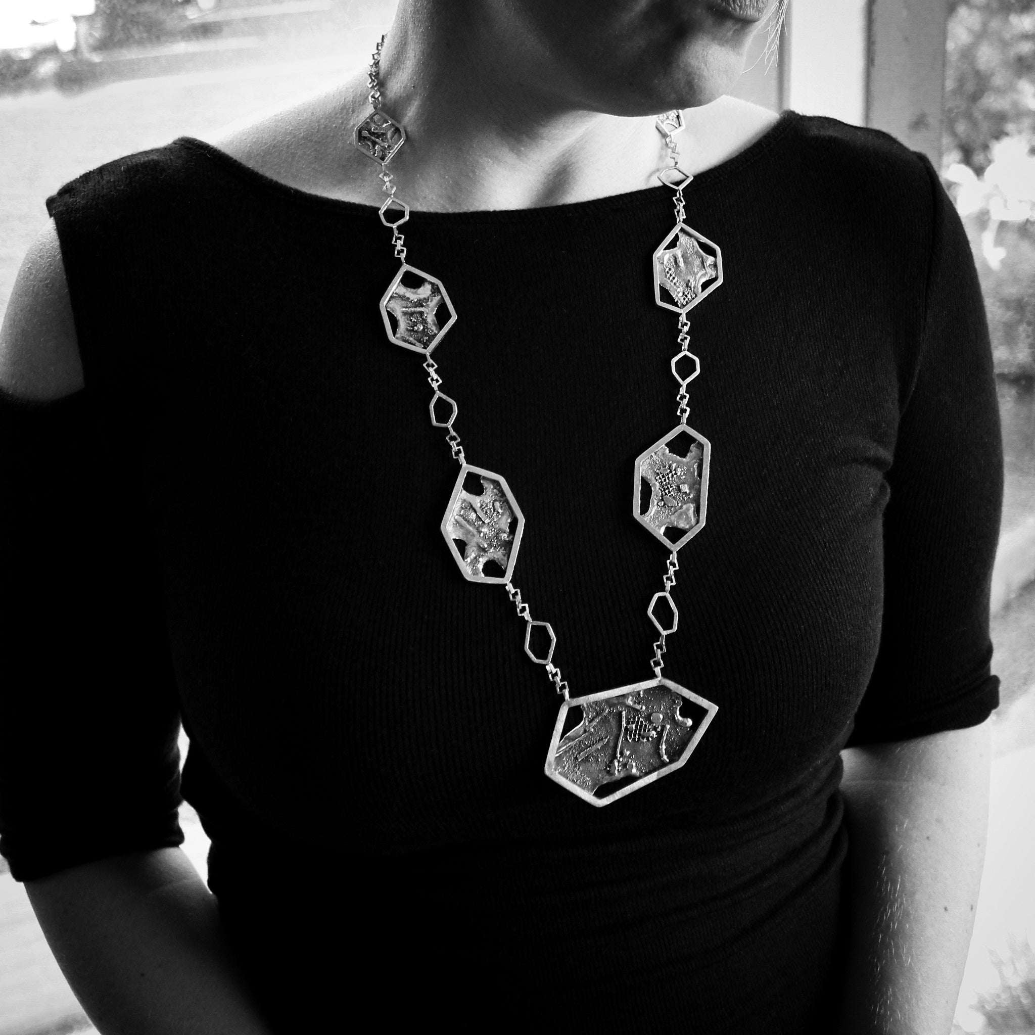 A close-up of woman’s torso and lower part of face, she looking towards left. She is wearing a black shirt, and a contemporary brutalist inspired necklace, with 7 asymmetric hexagons. Each hexagon is framing a textured back plate with melted holes. The hexagons are attached along a square link chain. Blurred trees can be seen in background. Image is black and white.