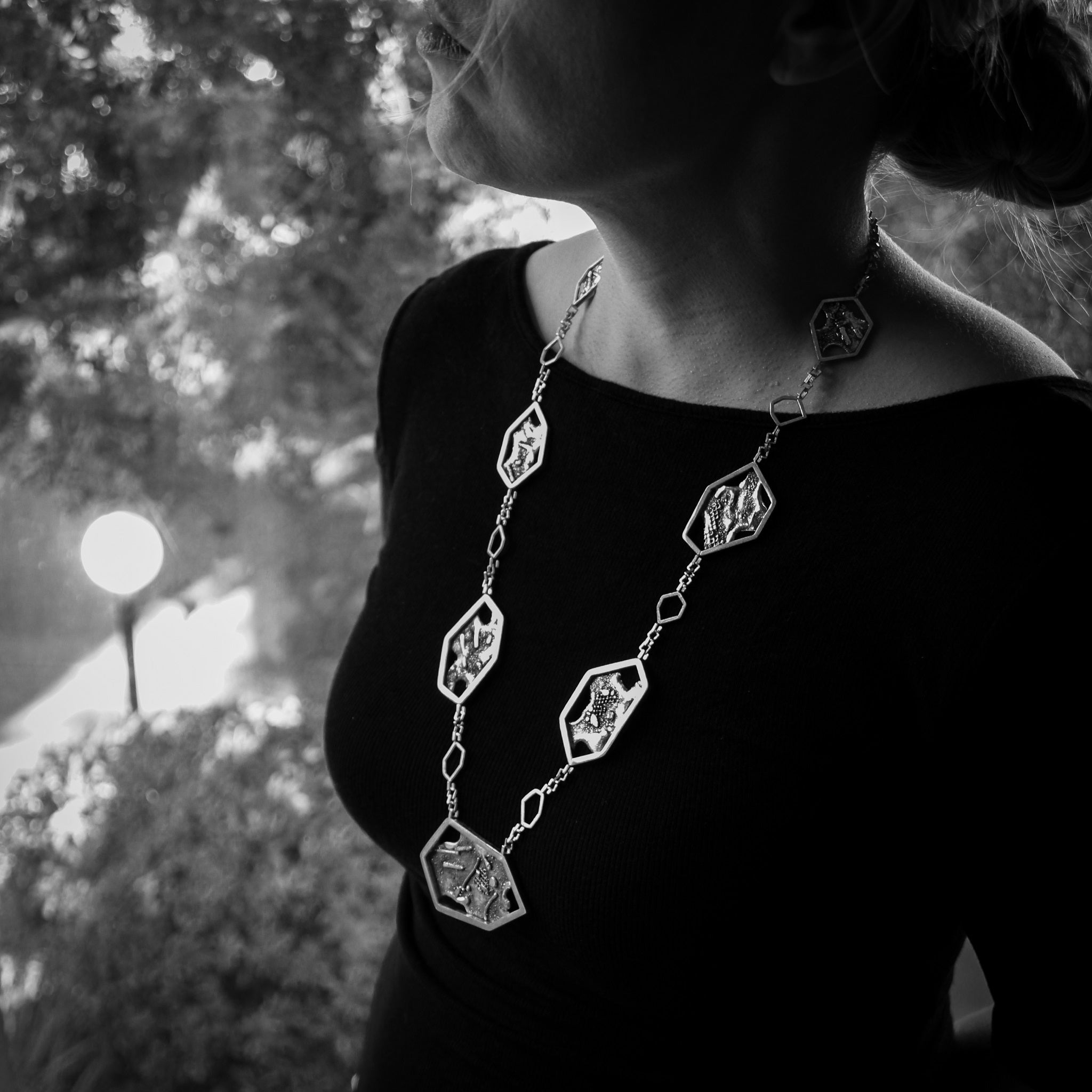 A close-up of woman’s torso and lower part of face, she looking towards right. She is wearing a black shirt, and a contemporary brutalist inspired necklace, with 7 asymmetric hexagons. Each hexagon is framing a textured back plate with melted holes. The hexagons are attached along a square link chain. A street light and trees are blurred and can be seen in background. Image is black and white.