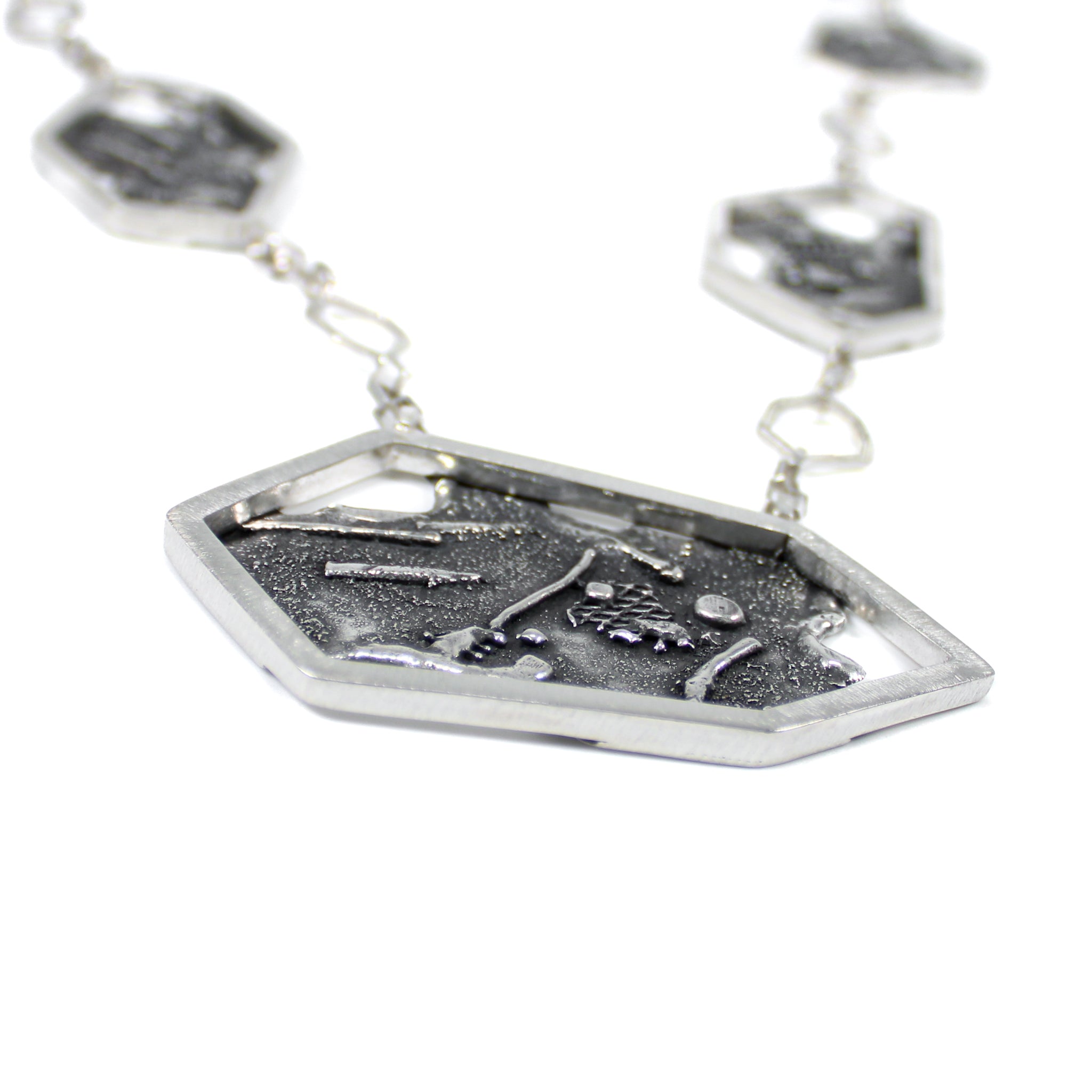 A necklace, laying flat. It is a close-up view of the side of the pendant, showing the edge of a hexagonal frame, highlighting the scratched texture on it. There are 3 other smaller hexagons on a square link chain, blurred in the background.  