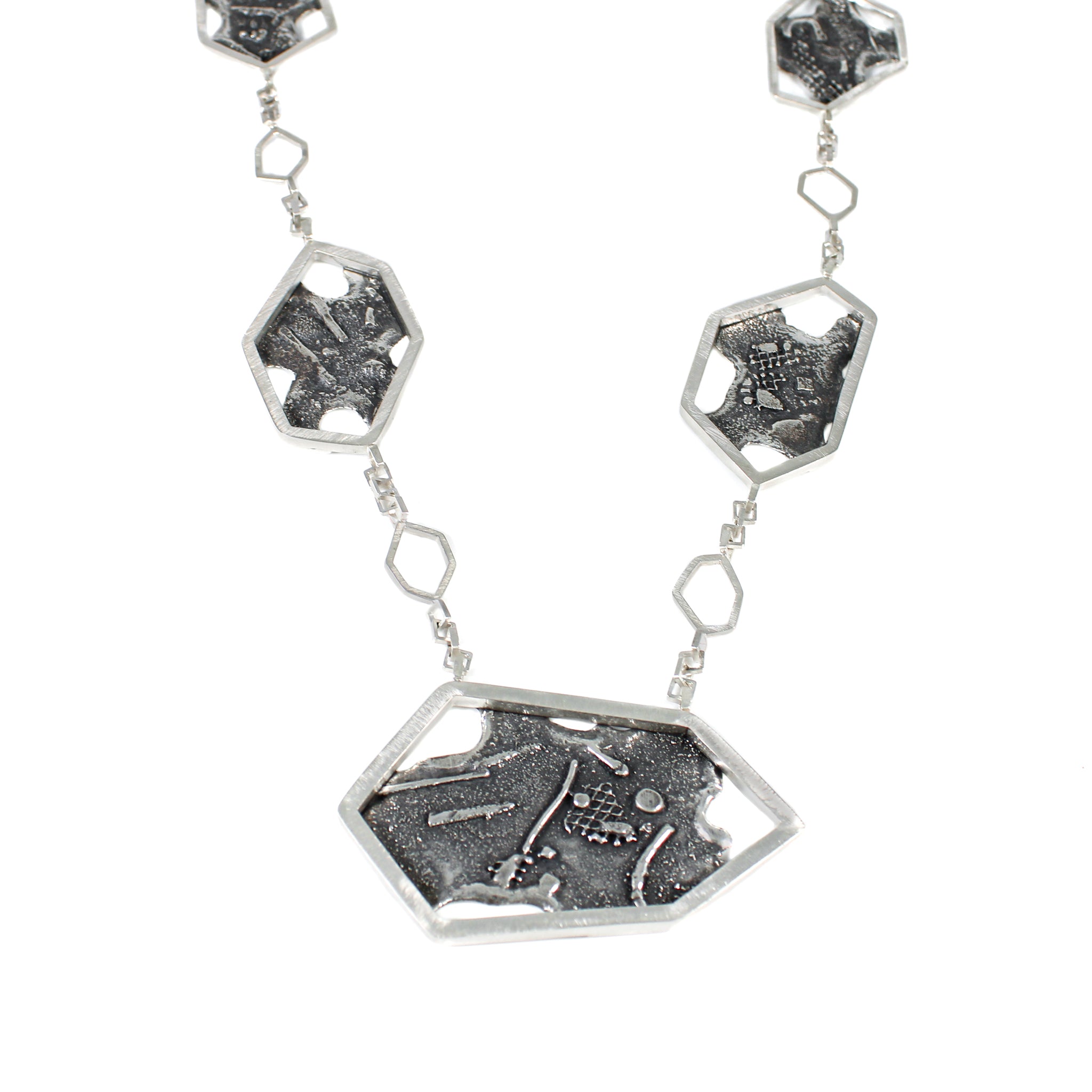 Front view of a modern sterling silver pendant, featuring a large central asymmetric, hexagon framing a fused and brutalist textured blackplate with melted holes. There are 2 more hexagons of descending size on either side, separated by an inch of square link chain with a smaller, hexagonal link in the middle.