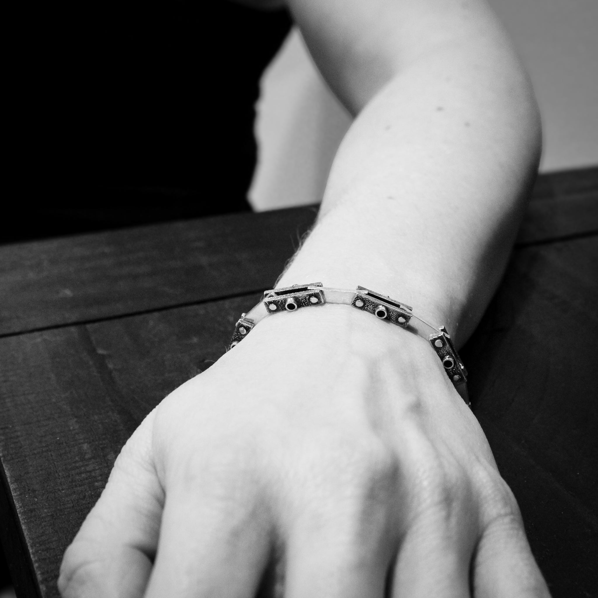 Black and white close-up of an arm resting on a black surface. The view is across the arm, highlighting the handmade silver bicycle chain bracelet.