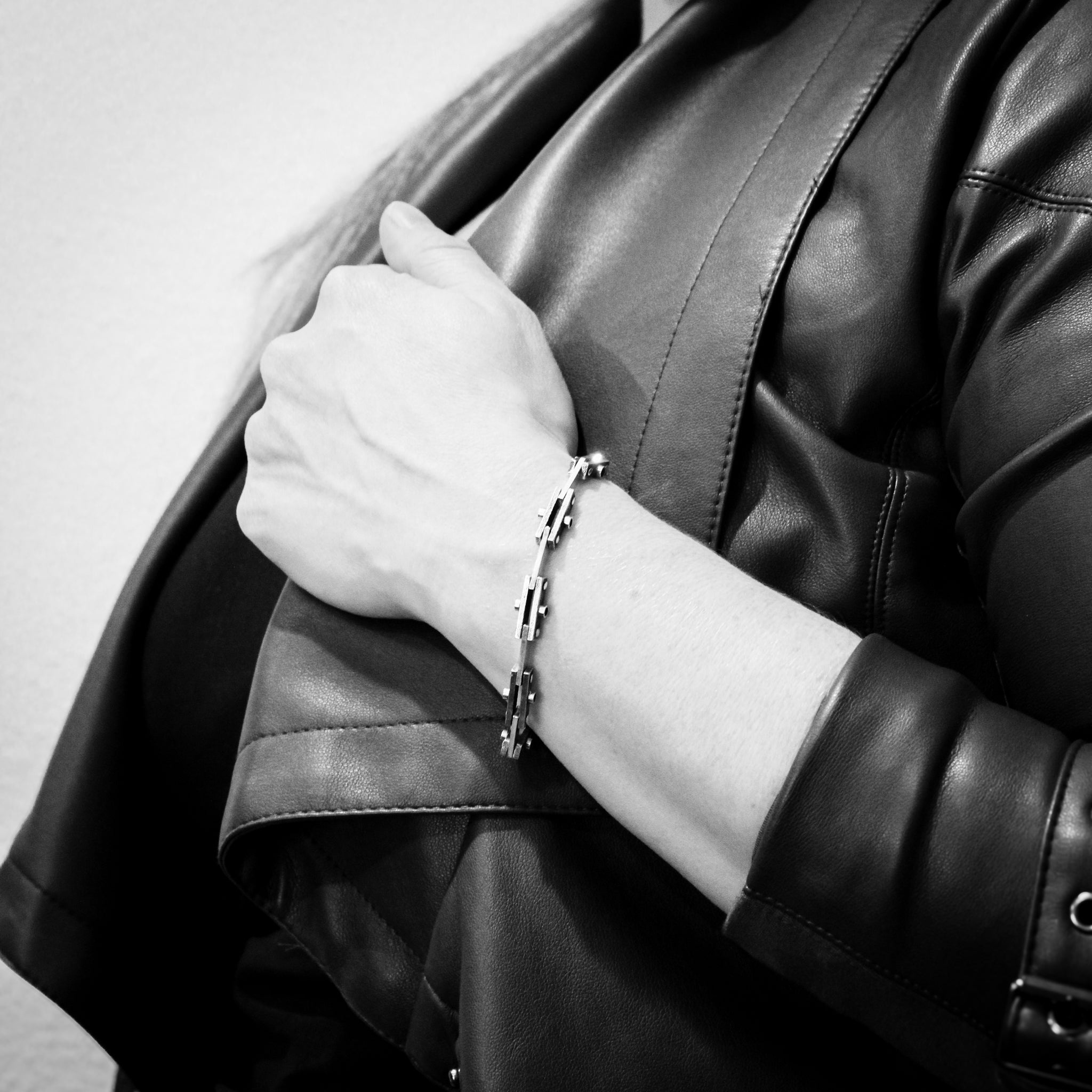 Black and white close-up of a woman's torso in a black leather jacket. Her arm is holding the edge of the jacket, and the image focuses on the sterling silver bicycle chain bracelet she is wearing