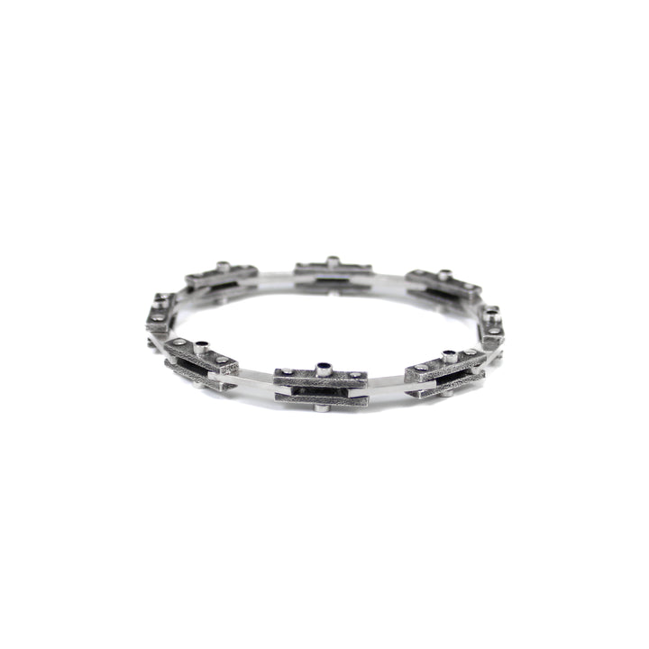 Unique industrial sterling silver bicycle-style chain bracelet. Every other link features a textured black finish with a 3mm tube soldered in the center on both sides and the alternate link is smooth and polished. Each link is connected by roughly finished rivets. Bracelet is closed and positioned in a circle on a white background. 