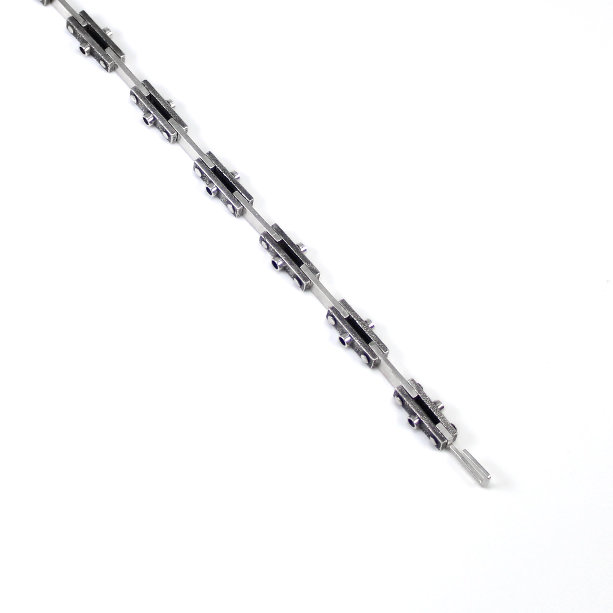 Unique industrial sterling silver bicycle-style chain bracelet. Every other link features a textured black finish with a 3mm tube soldered in the center on both sides and the alternate link is smooth and polished. Each link is connected by roughly finished rivets. Top down view of undone bracelet, laying flat across the background, highlighting a top view of the links and hook clasp. 