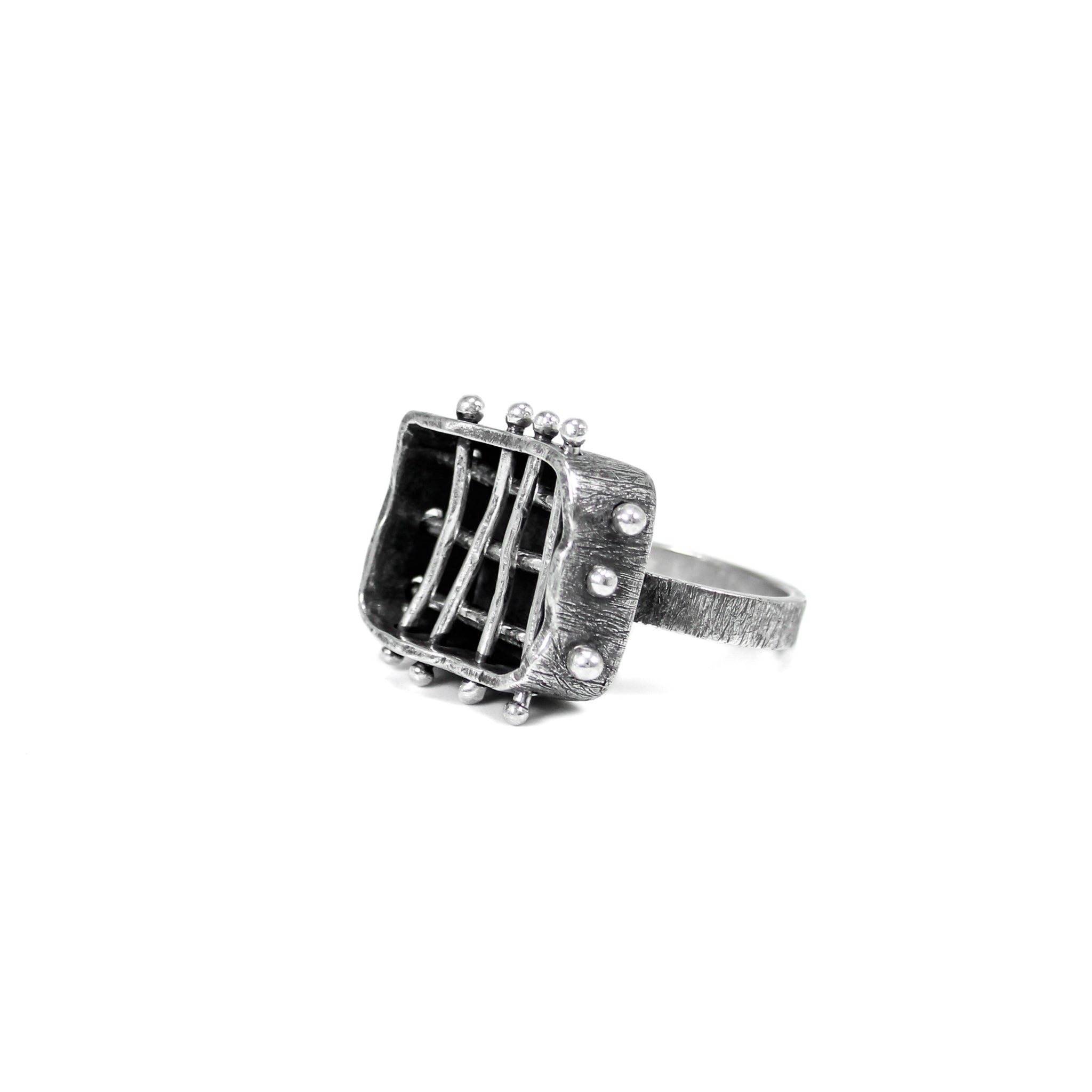 Forty-five-degree angle view of a brutalist inspired ring featuring a roughly crafted rectangle frame with three horizontally and four vertically positioned silver bars, creating a cage-like structure. The outside of the cage features an aggressively scratched and tarnished surface..