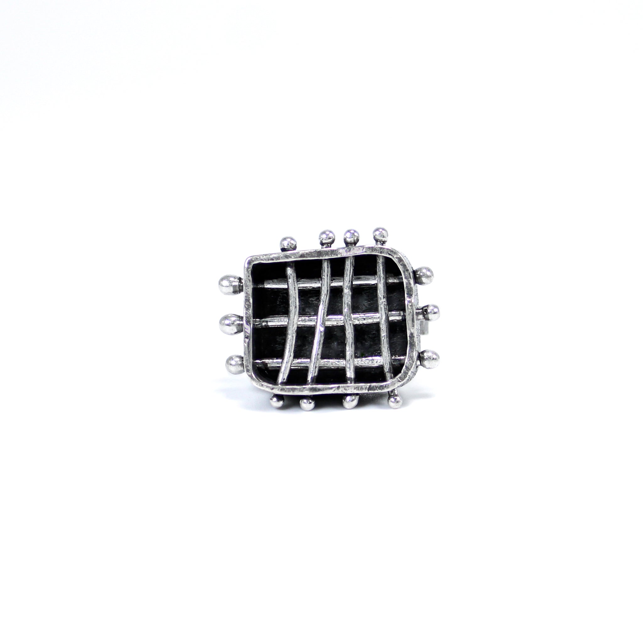 Front view of a brutalist inspired ring featuring a roughly crafted rectangle frame with three horizontally and four vertically positioned silver bars, creating a cage-like structure. The bars are intentionally bent and marred, piercing through the walls of the rectangle frame, with silver balls on either end holding them in place. The back of the cage is entirely black, providing a striking contrast