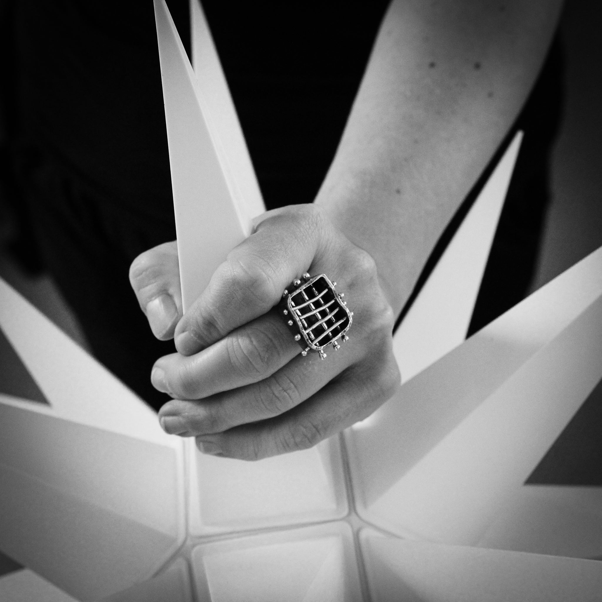 Black and white close-up of a hand holding the top a bright white spikey star. The focal point is the brutalist handmade sterling cage ring worn on the middle finger of the right hand.