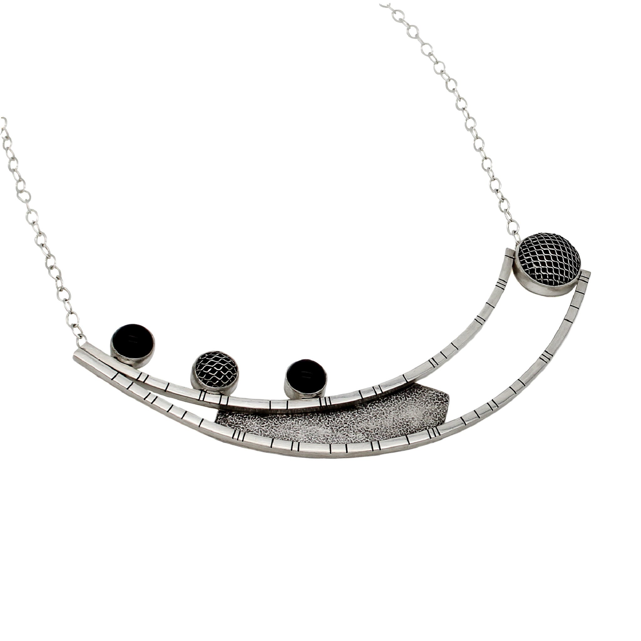 A necklace, laying flat, crafted from two long curved square wires with random black lines, connected at one end and split at the other. An angular sheet of blackened textured silver is positioned between the wires. On the right side of the top curved bar, three 10-millimeter stones are set – two black onyx and one silver mesh dome. On the left side, suspended between the ends of the curved bars, is a large bezel made of domed silver mesh. The pendant is attached to a link-in-link chain.