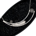 Load image into Gallery viewer, A necklace in a black dish filled with black pebbles. The pendant is fabricated from two long curved square wires with random black lines, connected at one end and split at the other. A sheet of blackened textured silver is positioned between the wires. 3 stones are set on the right side of pendant - 2 onyx and 1 domed mesh silver.  On the left side, between the ends of the curved bars, is a large bezel made of domed silver mesh
