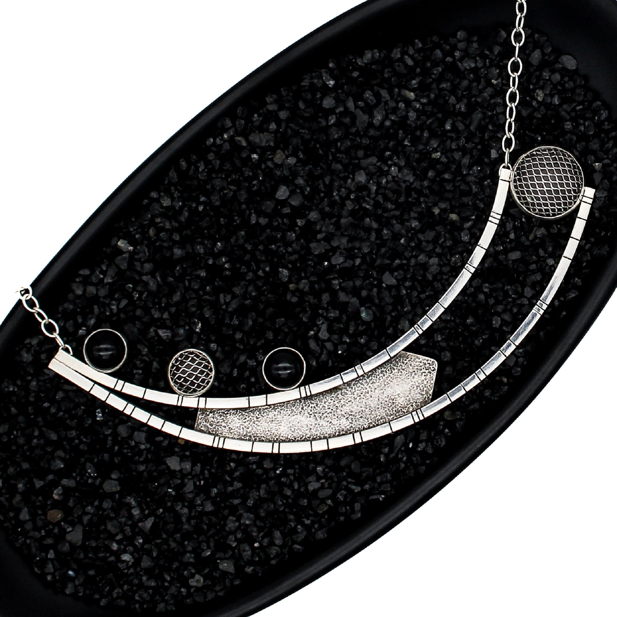 A necklace in a black dish filled with black pebbles. The pendant is fabricated from two long curved square wires with random black lines, connected at one end and split at the other. A sheet of blackened textured silver is positioned between the wires. 3 stones are set on the right side of pendant - 2 onyx and 1 domed mesh silver.  On the left side, between the ends of the curved bars, is a large bezel made of domed silver mesh