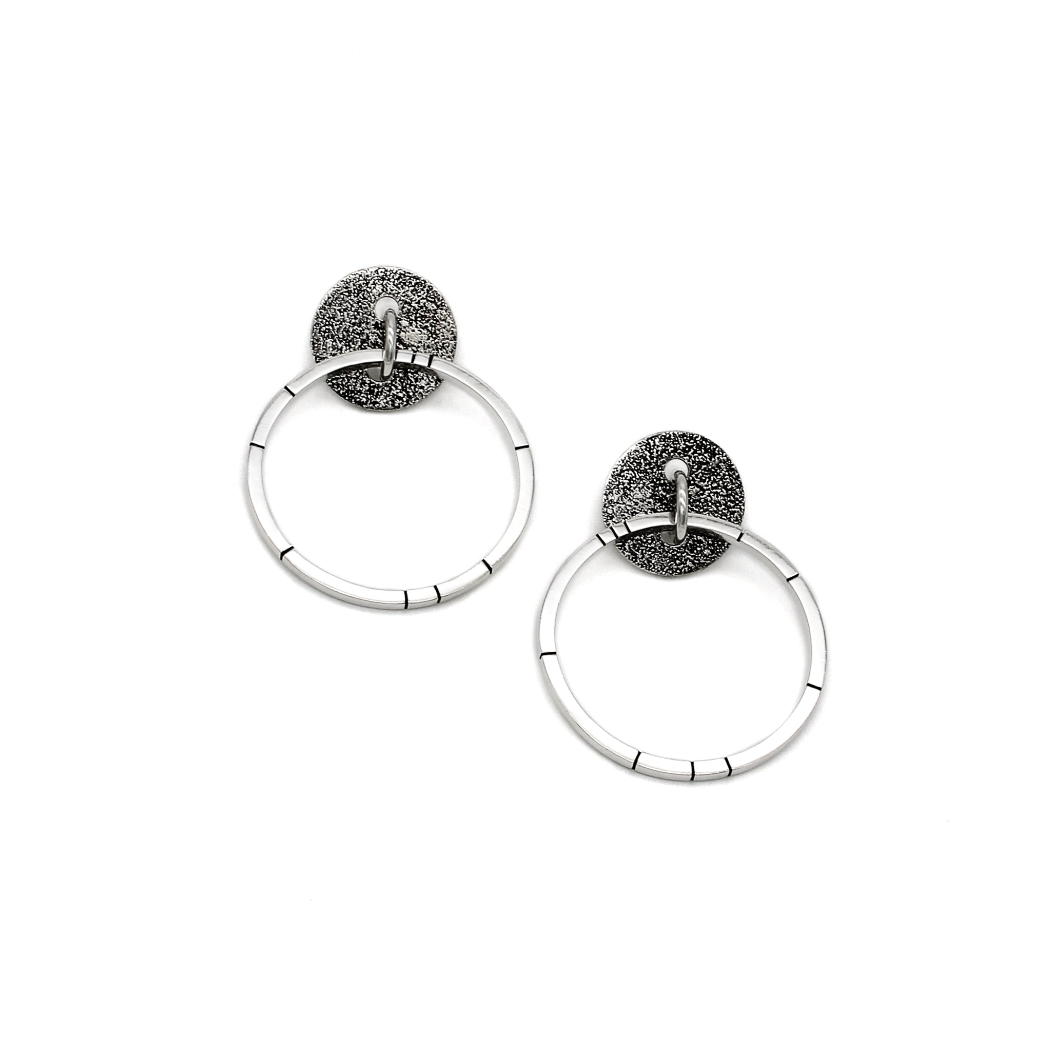 Unique handmade earrings with a blackened, round, brutalist-textured disc. A ring runs through the center, and a larger hoop hangs through the ring, mimicking a door knocker. Random black lines accentuate the face of the hoop for added character.