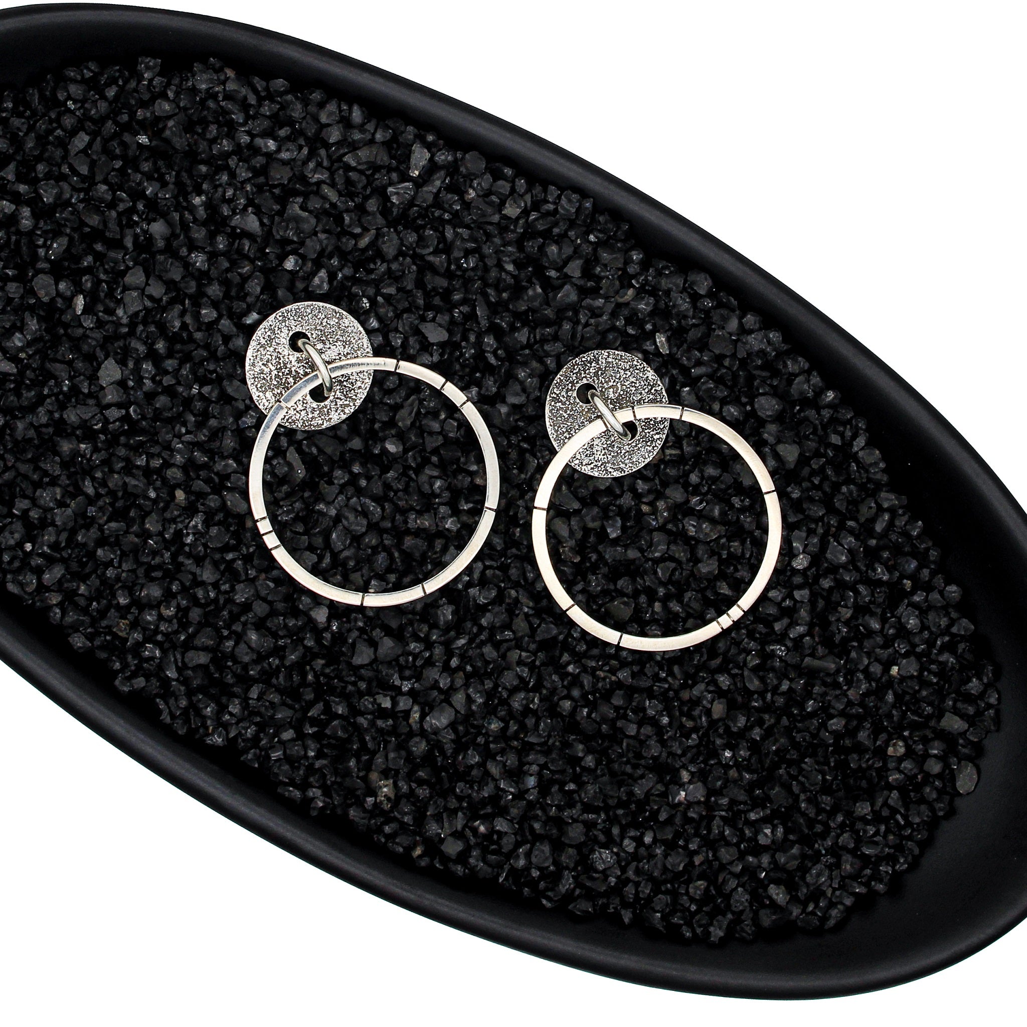 Unique handmade earrings with a blackened, round, brutalist-textured disc. A ring runs through the center, and a larger hoop hangs through the ring, mimicking a door knocker. Random black lines accentuate the face of the hoop for added character. Earrings are in a black dish, with black pebbles, on a white background.