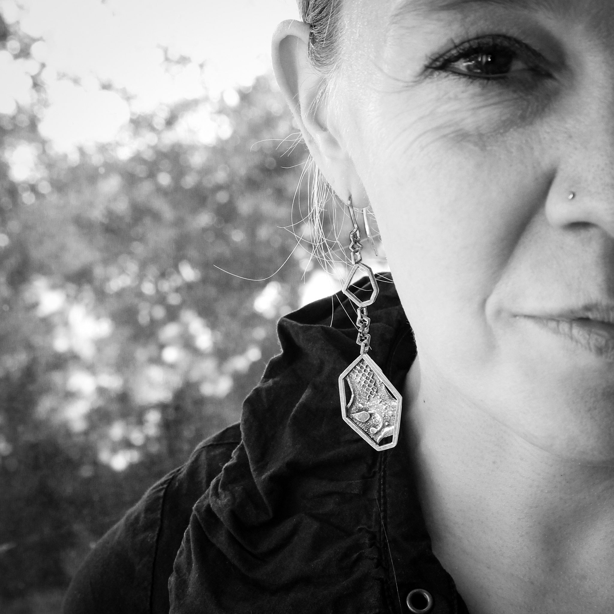 Black and white close-up on right side of woman’s face that is bisected by right edge of photo. She is wearing a black jacket with a large collar. The center of photo is focused on the unique sterling hexagonal, brutalist inspired earring she is wearing. Blurred trees can be seen in the background.