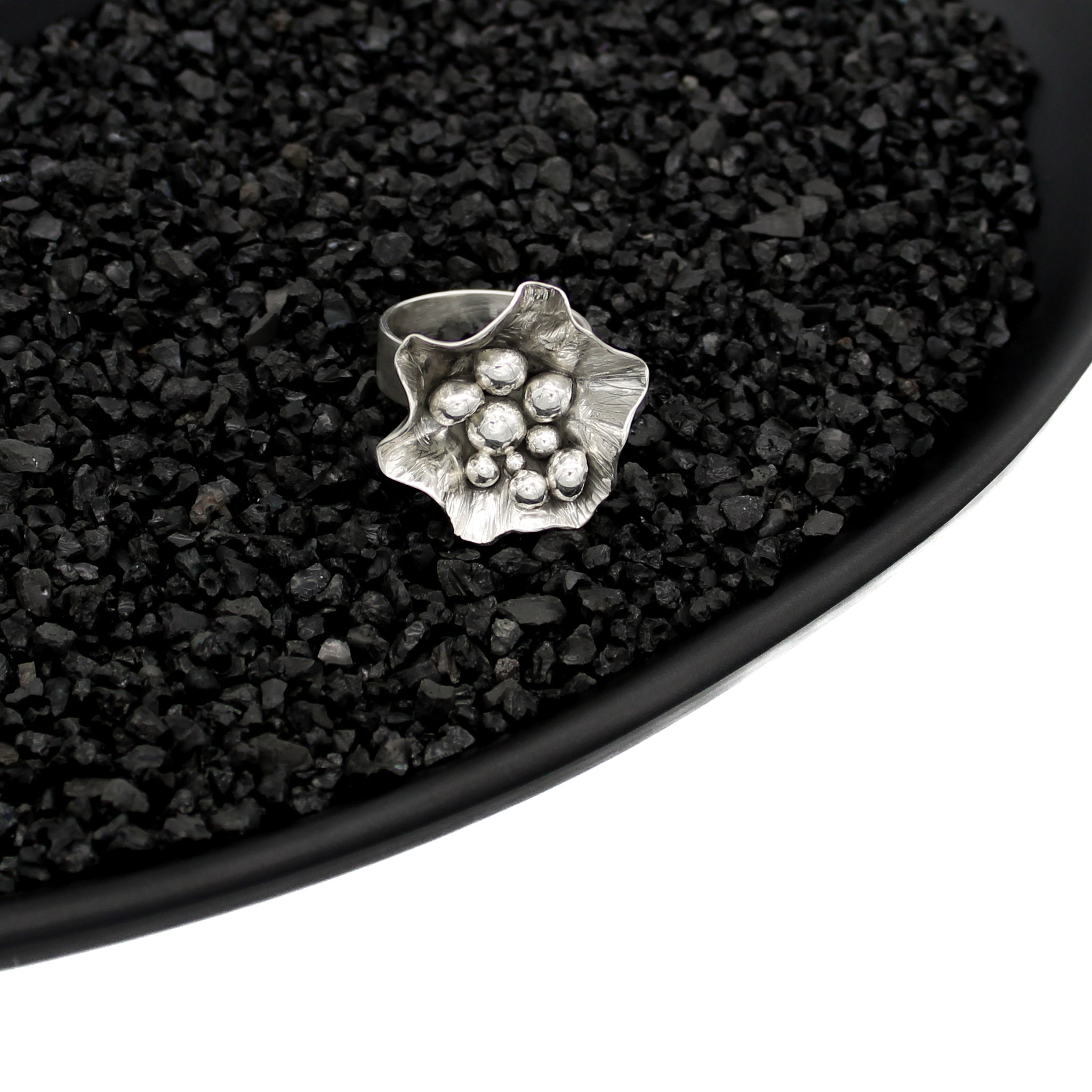 Sterling silver, handmade, freeform flower ring filled with several asymmetric varying sized balls, in a black dish filled with black pebbles on white background. 