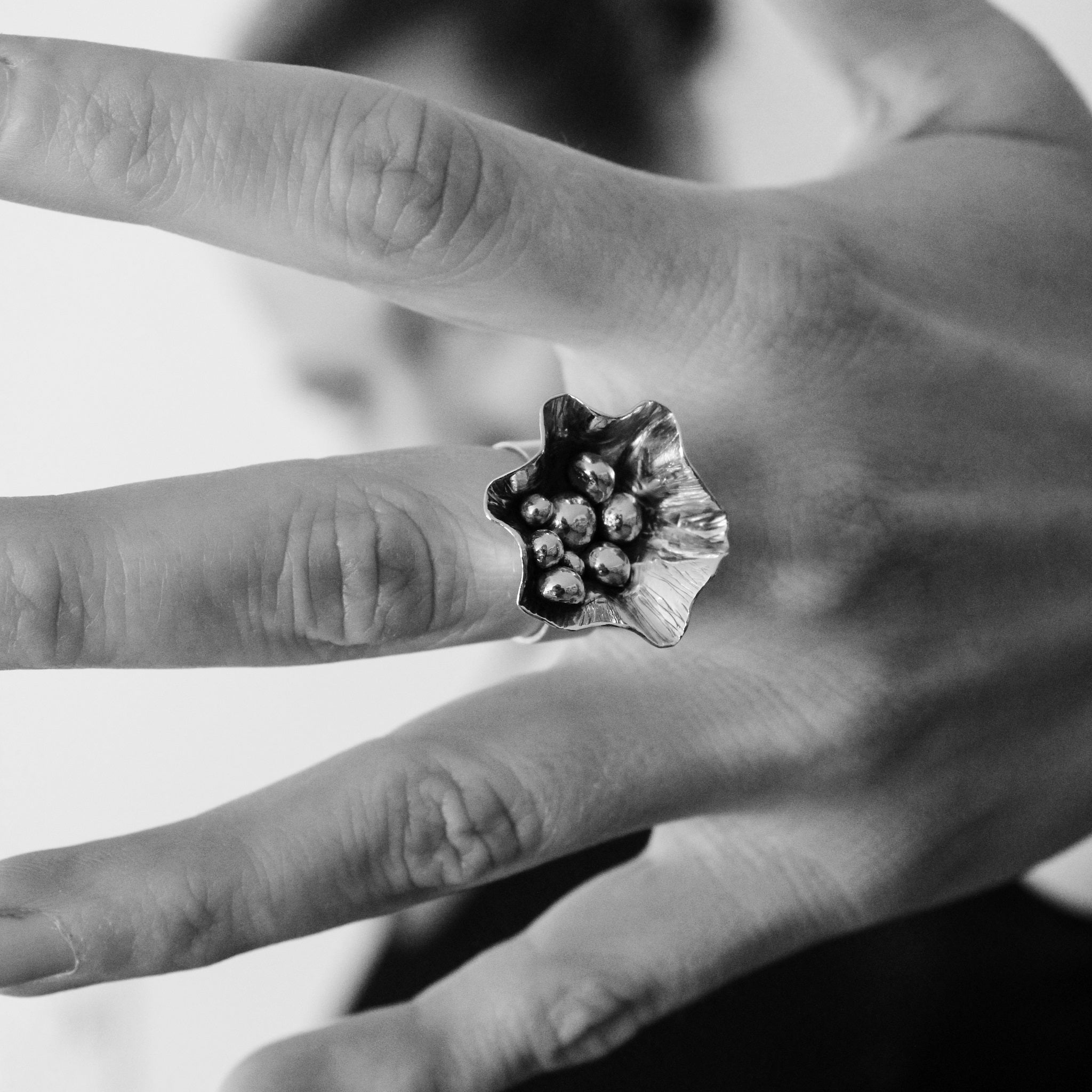 Black and white close up of unique, handmade, sterling silver flower ring on hand with spread fingers. A woman's blurred face in seen in background between fingers. 